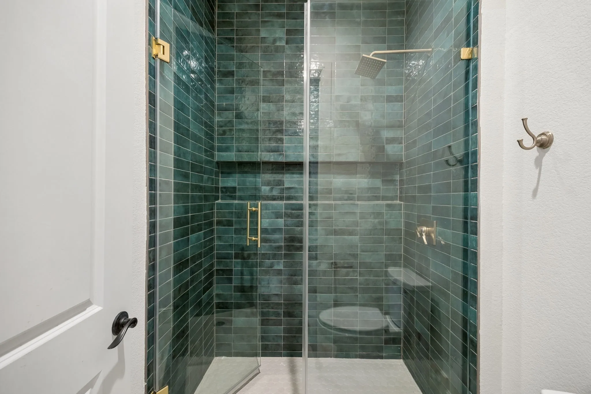 Walk-in shower with emerald green subway tile, frameless glass enclosure, built-in recessed shelf, and gold-finish hardware