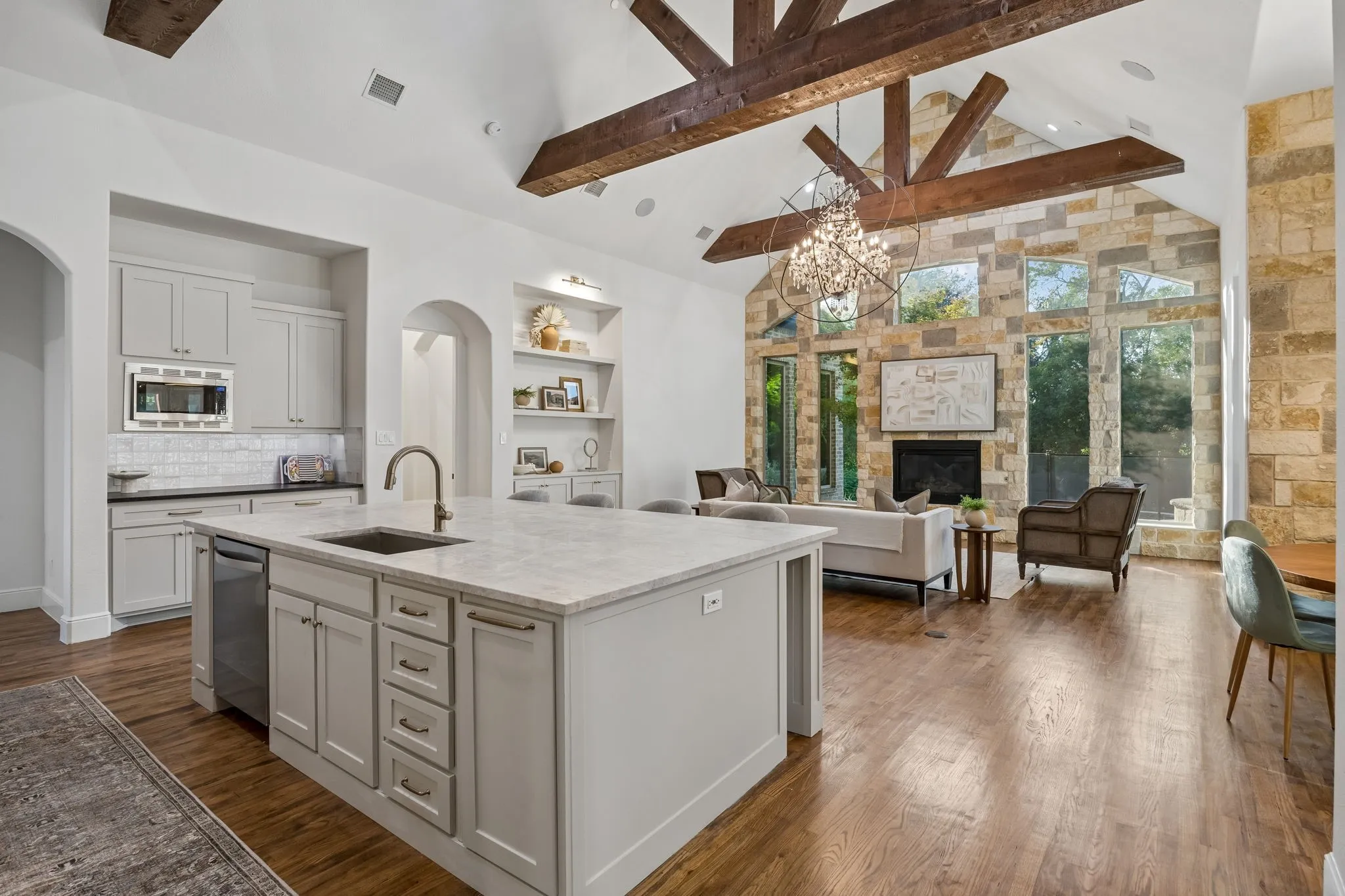 Spacious open-concept living area featuring a large kitchen island with integrated sink, vaulted ceilings with exposed wood beams, a stone accent wall with a fireplace, and extensive window-framing