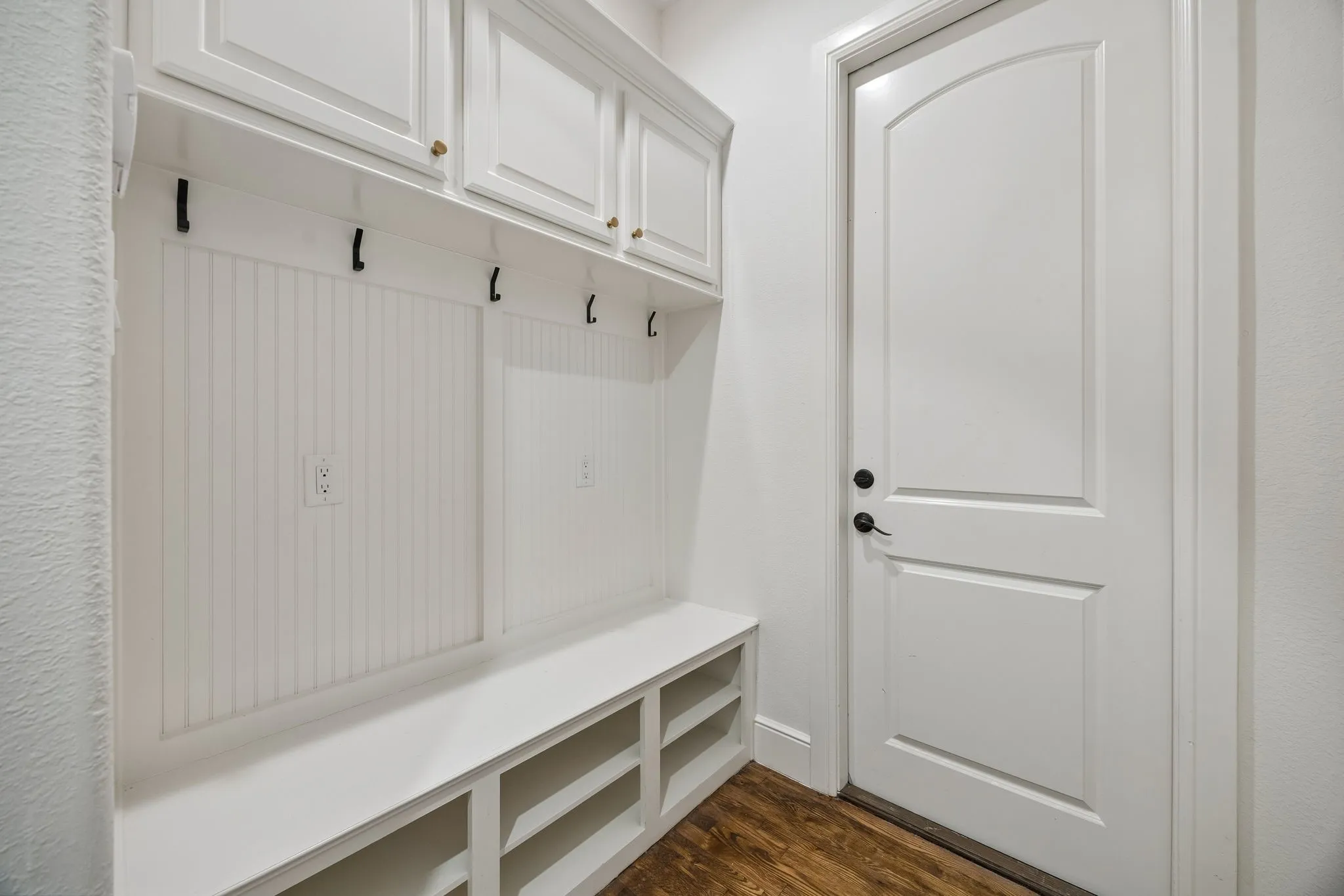 Built-in storage bench with cubbies, beadboard paneling, upper cabinetry with gold-toned hardware, and dark-toned coat hooks