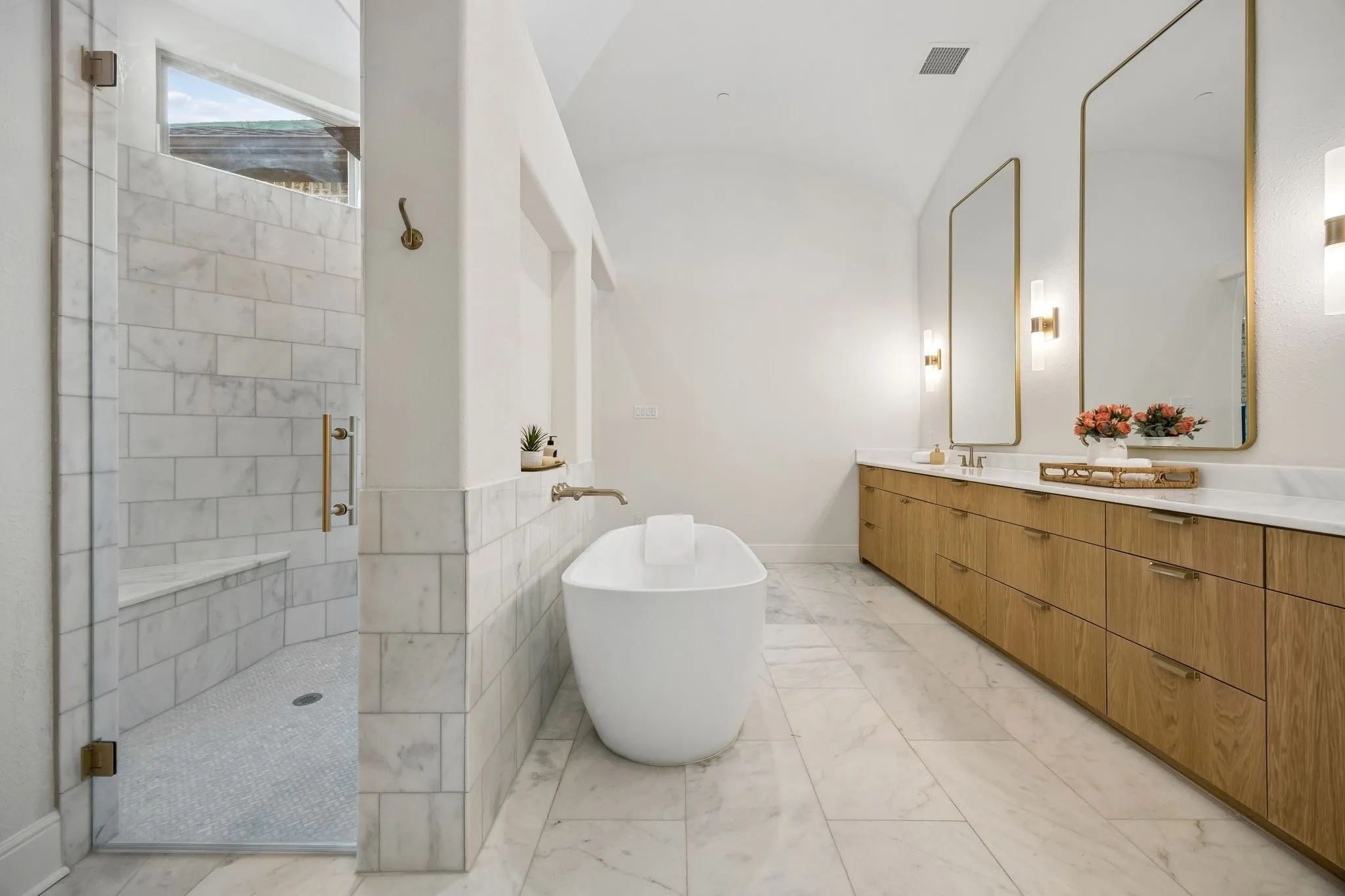 Spacious bathroom featuring a freestanding soaking tub, dual vanity with wood-finish cabinetry and rectangular mirrors, a walk-in shower with bench seating, and marble tile flooring