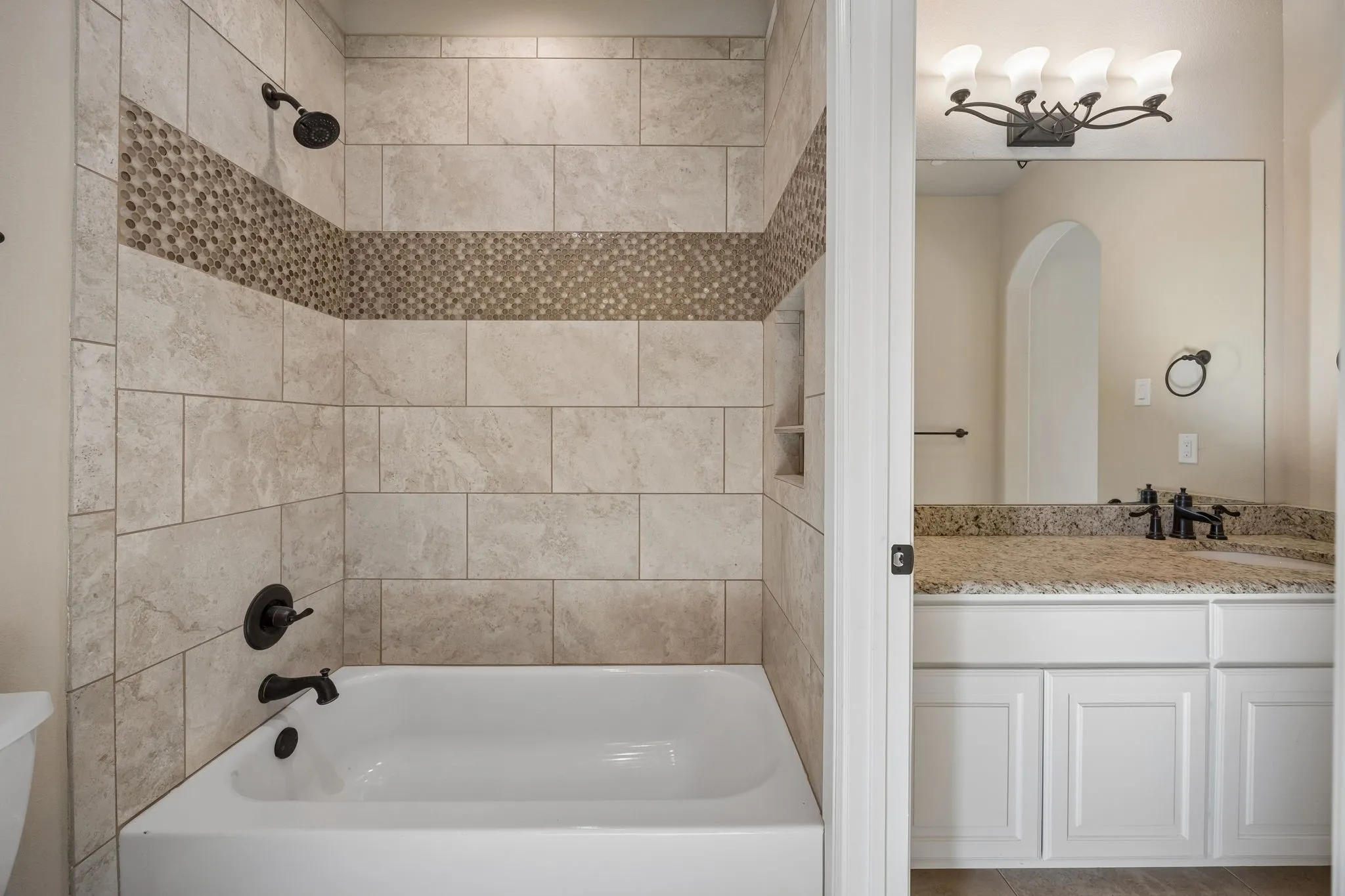 Bathroom featuring a tiled tub and shower surround with an accent mosaic band, oil-rubbed bronze fixtures, a built-in shower niche, a granite-finish vanity countertop, and white cabinetry