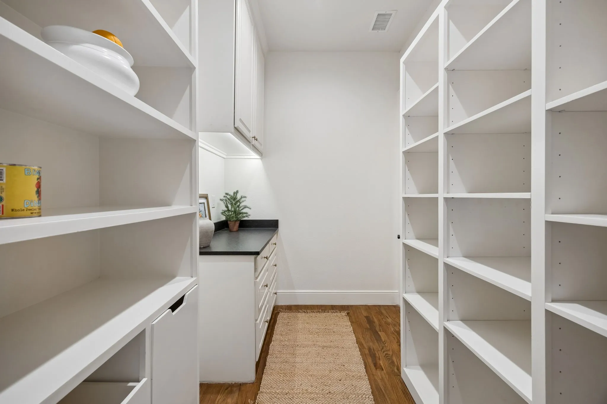 Dedicated pantry featuring extensive white shelving, integrated cabinetry, dark countertop space, and wood-finish flooring