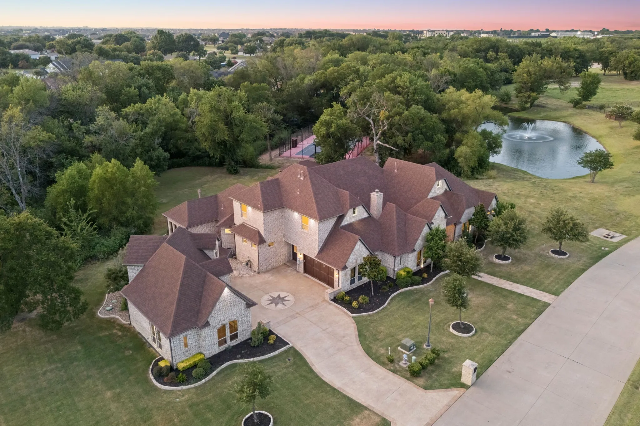 Expansive property featuring a multi-gabled residence with stone and stucco exterior, a private pond with a water feature, extensive landscaping with mature trees, a long driveway with a compass rose inlay, and an additional detached structure