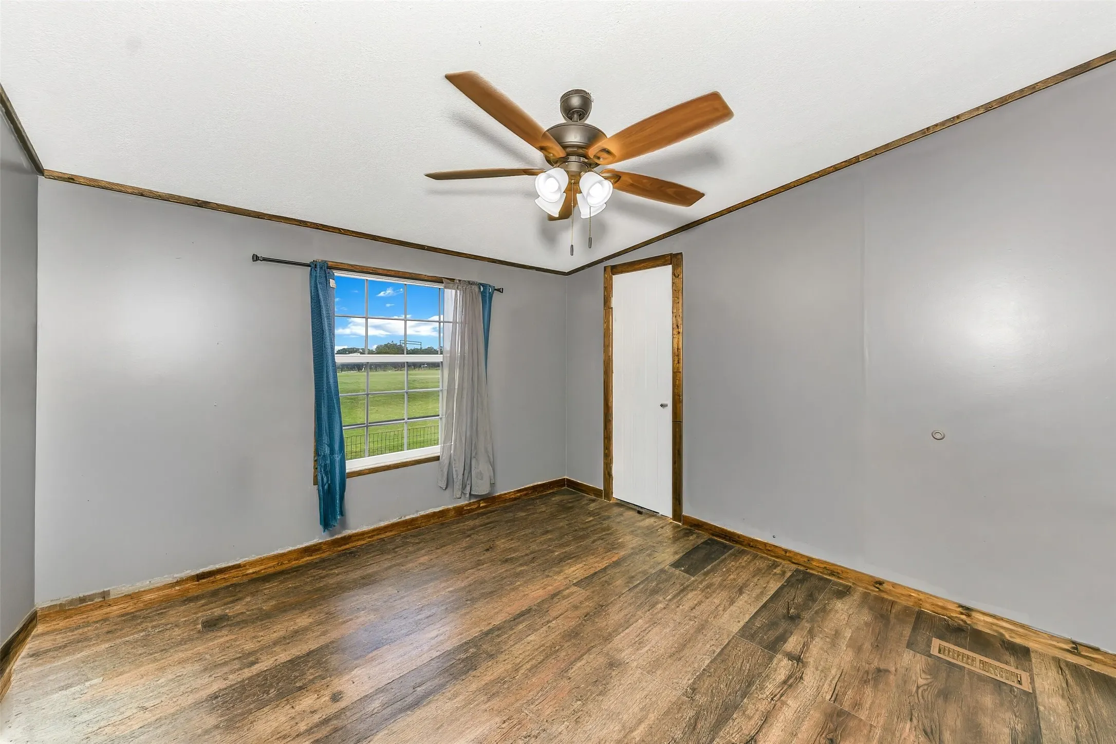Spacious room with wood-finish flooring, a ceiling fan with integrated lighting, and a window with outdoor scenery-AI removal of furniture
