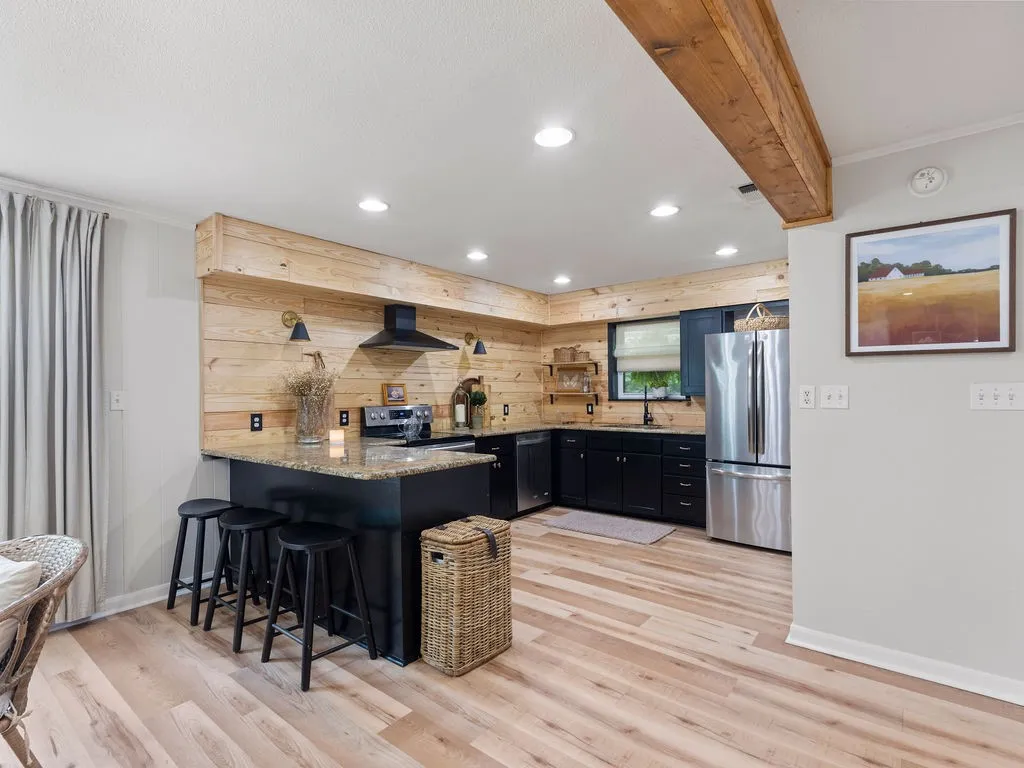 Kitchen featuring black cabinetry with stainless steel appliances, wood-finish flooring, a light-toned wooden plank accent wall, an island with a speckled countertop, and recessed lighting