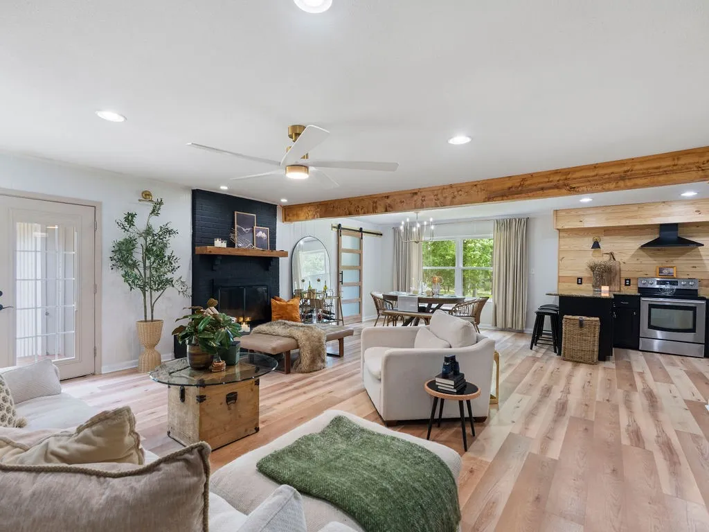 Spacious living area featuring light wood-finish flooring, a black brick fireplace with a wood mantle, and exposed beam accents