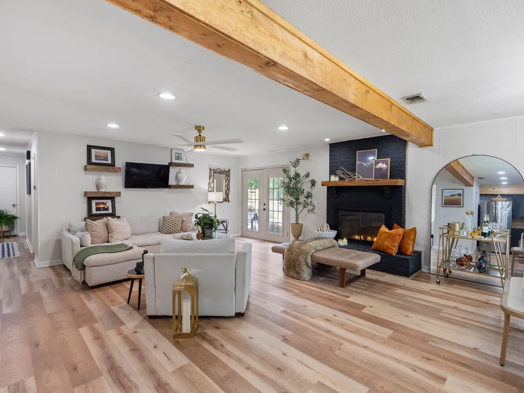 Spacious living area featuring wood-finish flooring, a black brick fireplace with a wood mantel, exposed ceiling beams, recessed lighting, and a ceiling fan