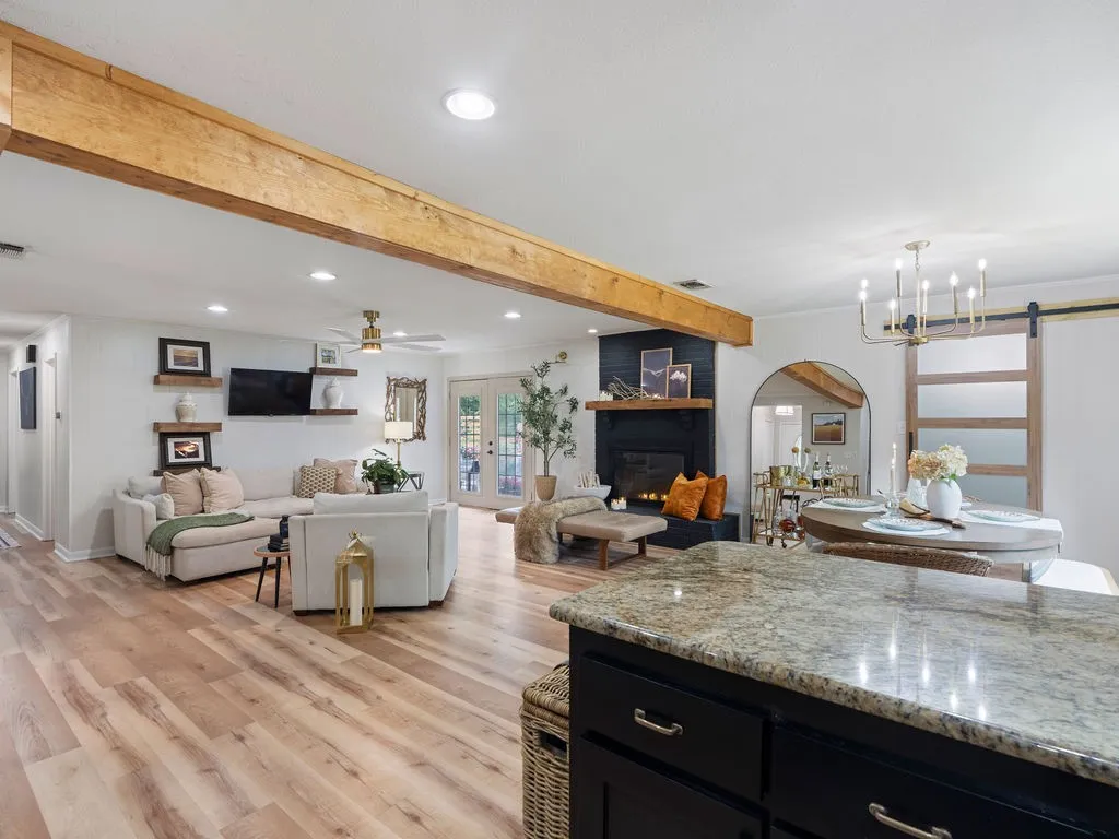 Open-concept living space featuring wood-finish flooring, exposed ceiling beams, a black-painted fireplace with a wood mantle, and a kitchen island with a light-toned countertop