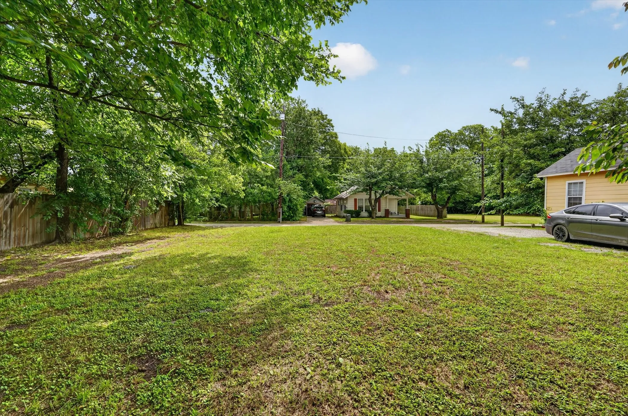 Expansive grassy lot bordered by a wooden fence, featuring mature shade trees and a gravel driveway