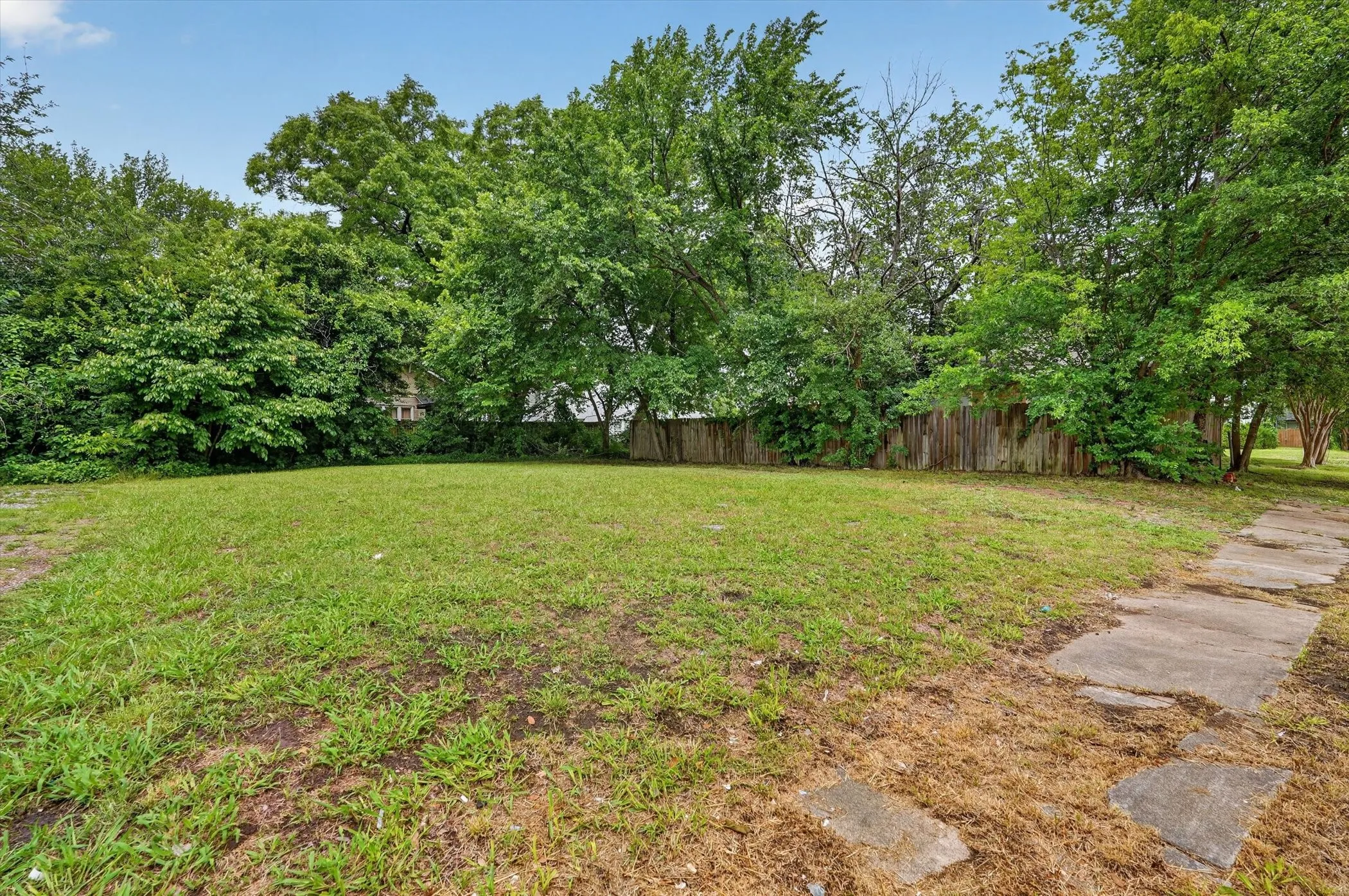 Expansive grass area with mature perimeter trees, a partial wood fence, and a concrete paver pathway