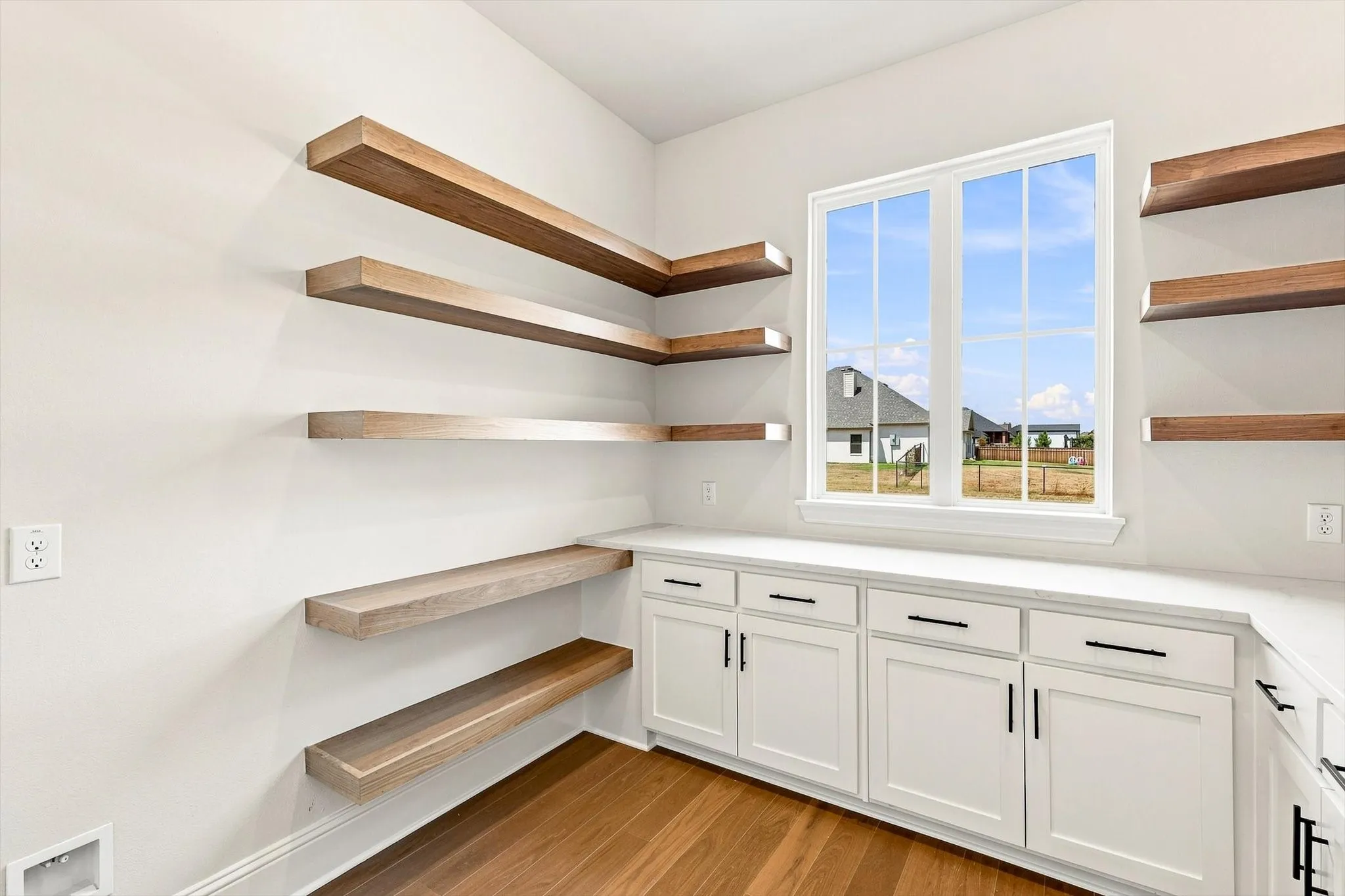 Expansive floating shelves, white cabinetry with black hardware, wood-finish flooring, and a large window