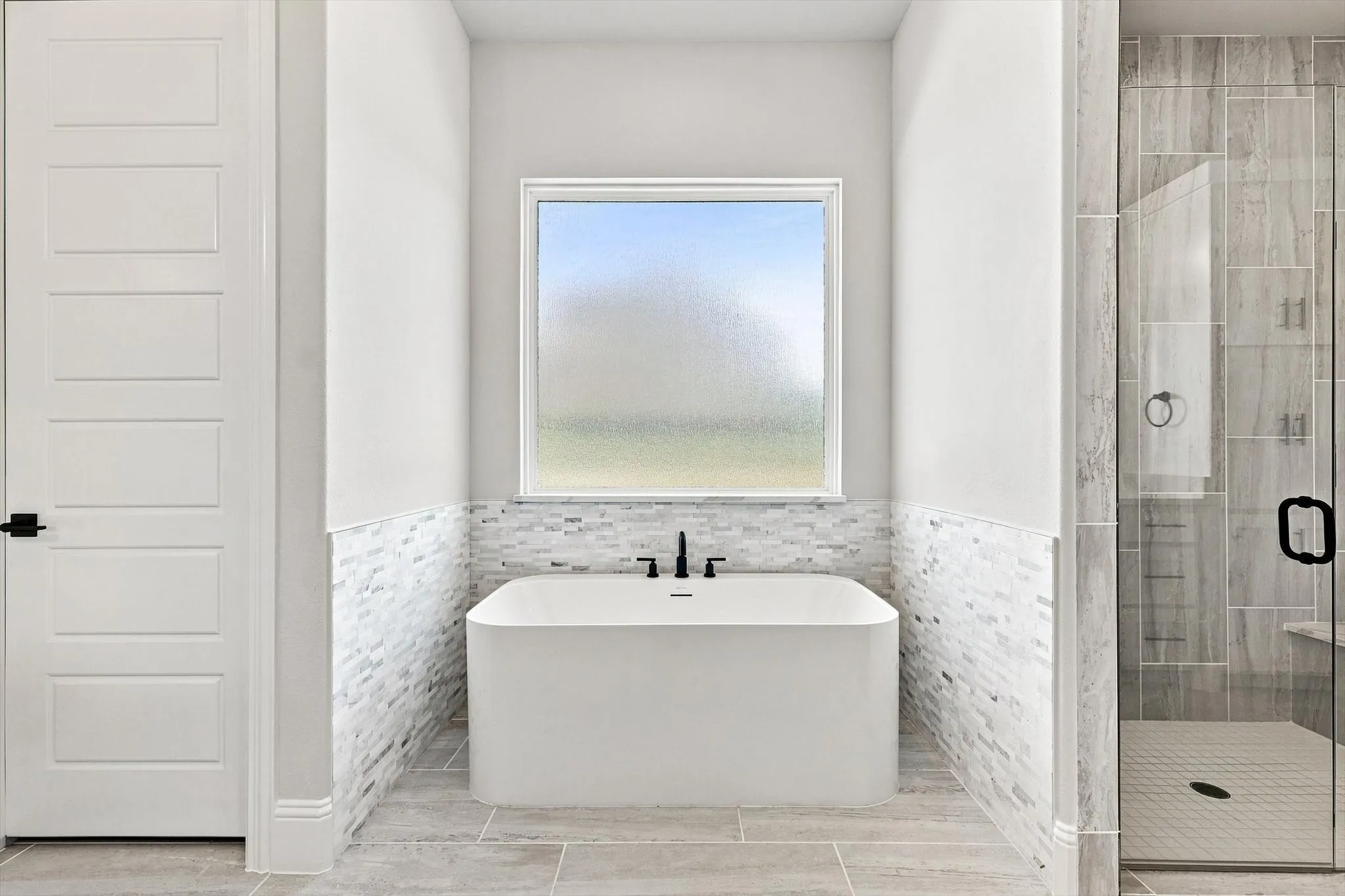 Freestanding soaking tub with matte black fixtures, framed window, tile wainscoting, and large format tile flooring