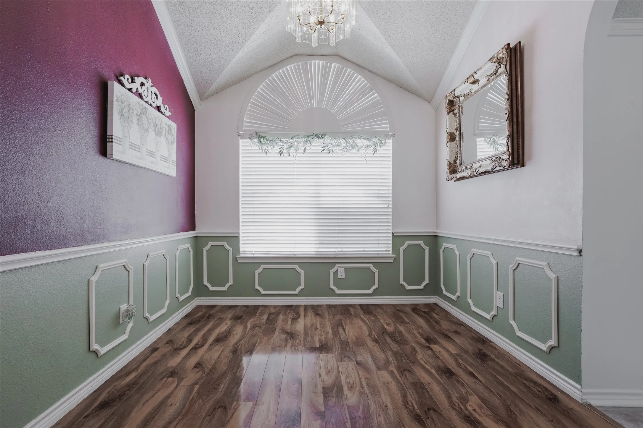 Elegant hallway showcasing architectural detail with vaulted ceiling, arched transom window with plantation shutters, warm wood-finish flooring, and wainscoting with decorative molding throughout