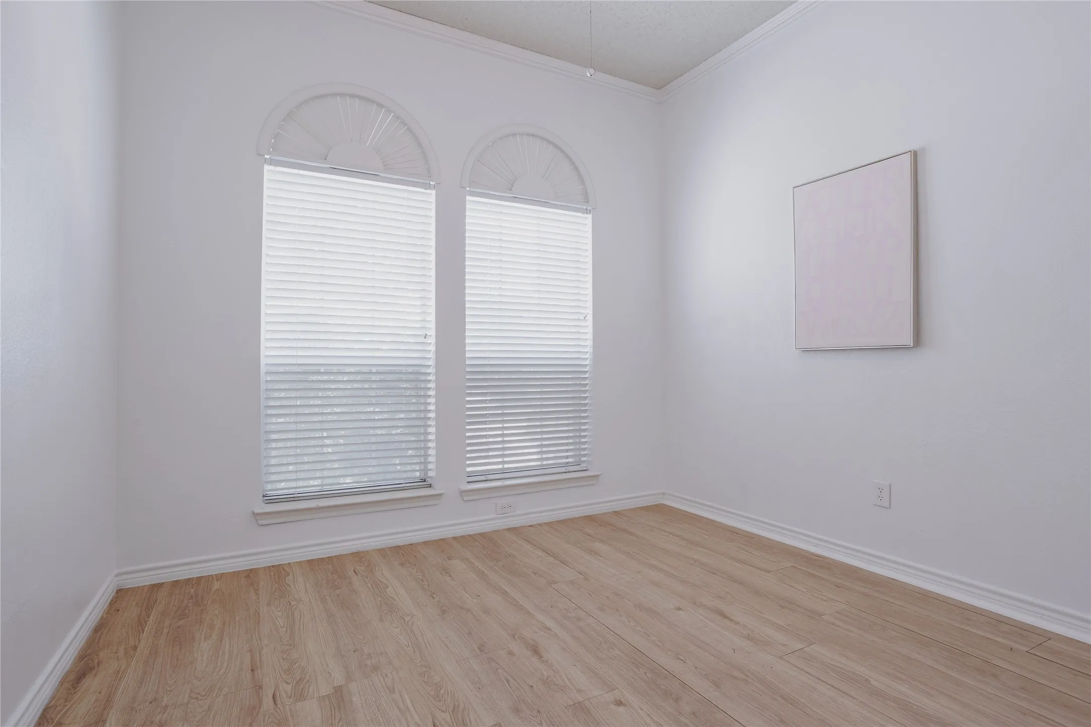 Bright corner room featuring arched transom windows with plantation shutters, warm wood-finish flooring, and clean white walls with crown molding detail