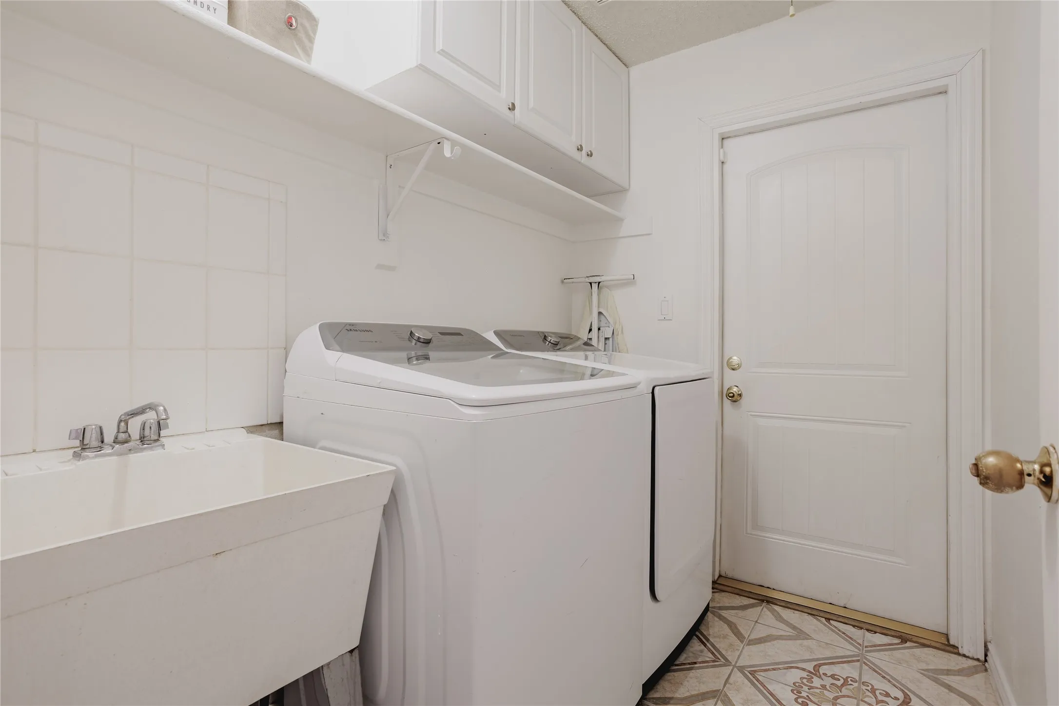 Dedicated laundry room featuring a top-load washer with dual wash basins, polished chrome faucetry, white subway tile walls, overhead cabinetry for storage, patterned tile flooring, and a white six-panel entry door