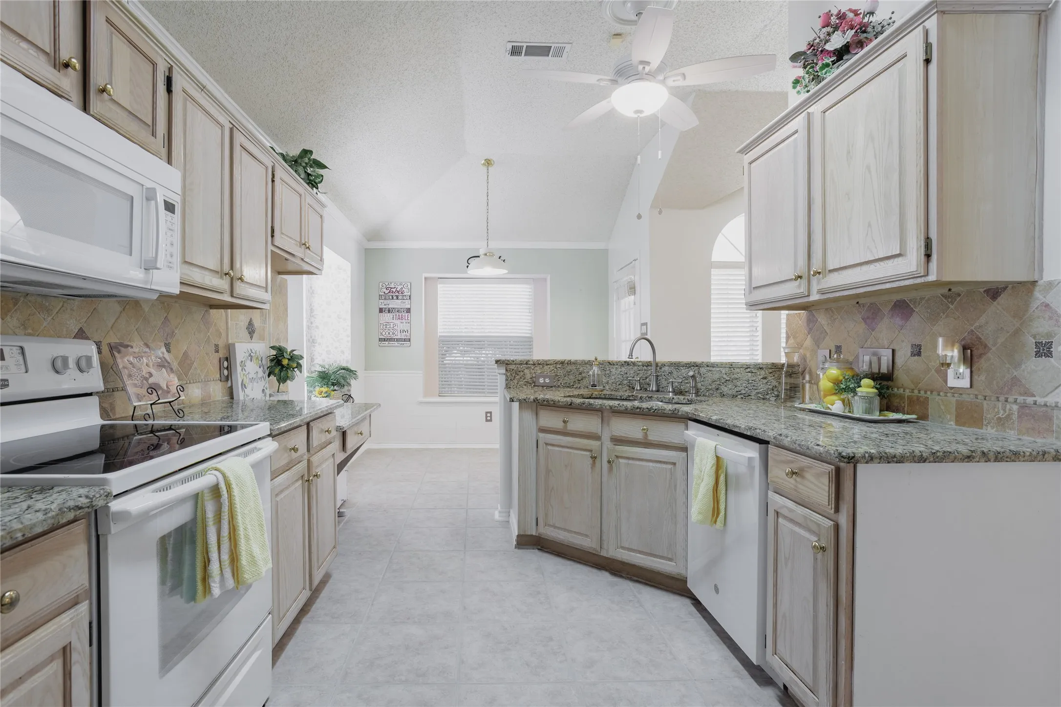 Galley kitchen featuring light wood-finish cabinetry, patterned tile backsplash, granite countertops, and built-in appliances