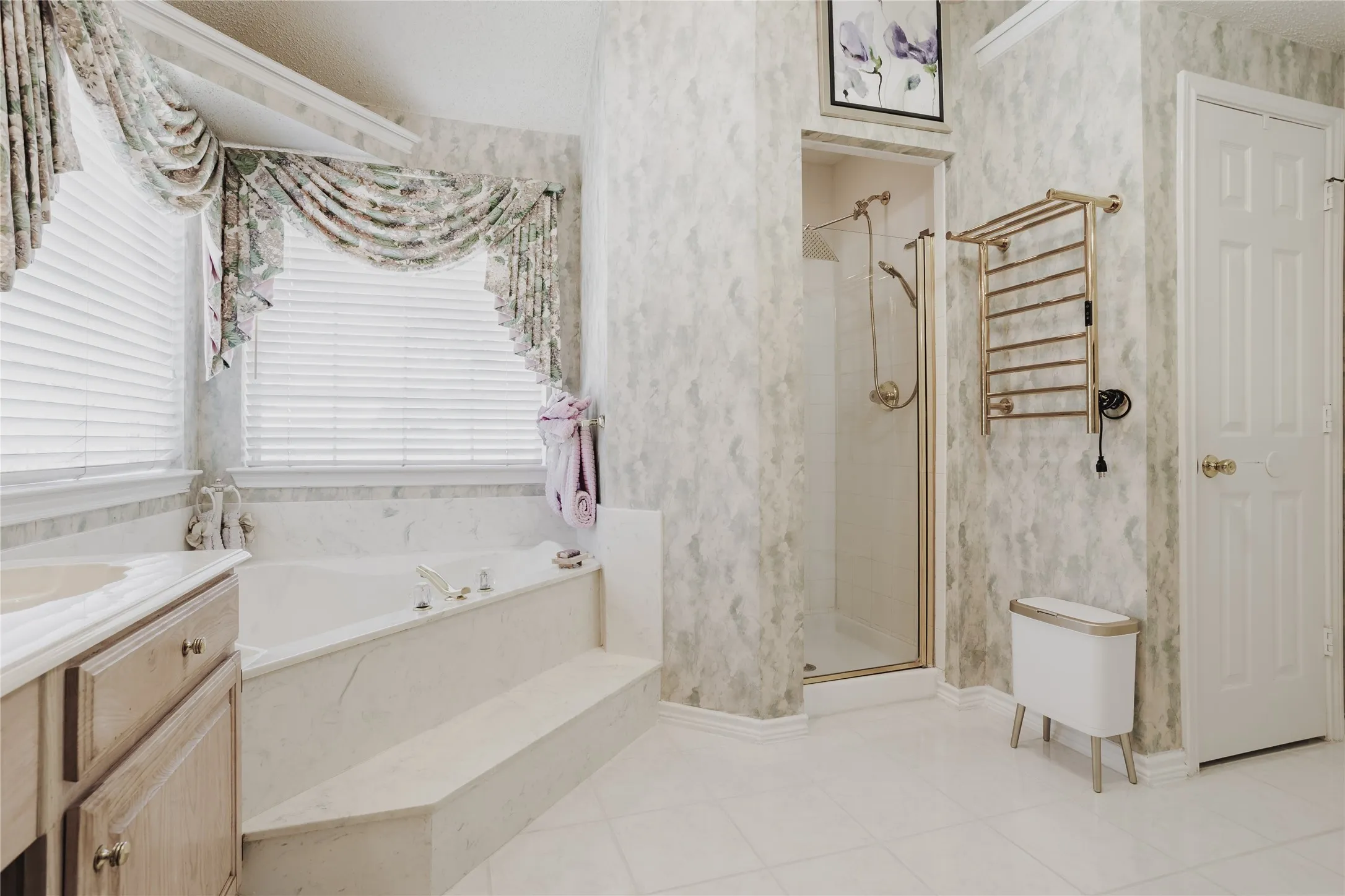 Built-in soaking tub with marble-look surround, walk-in shower with glass enclosure, white tile flooring, and light-toned vanity with integrated sink