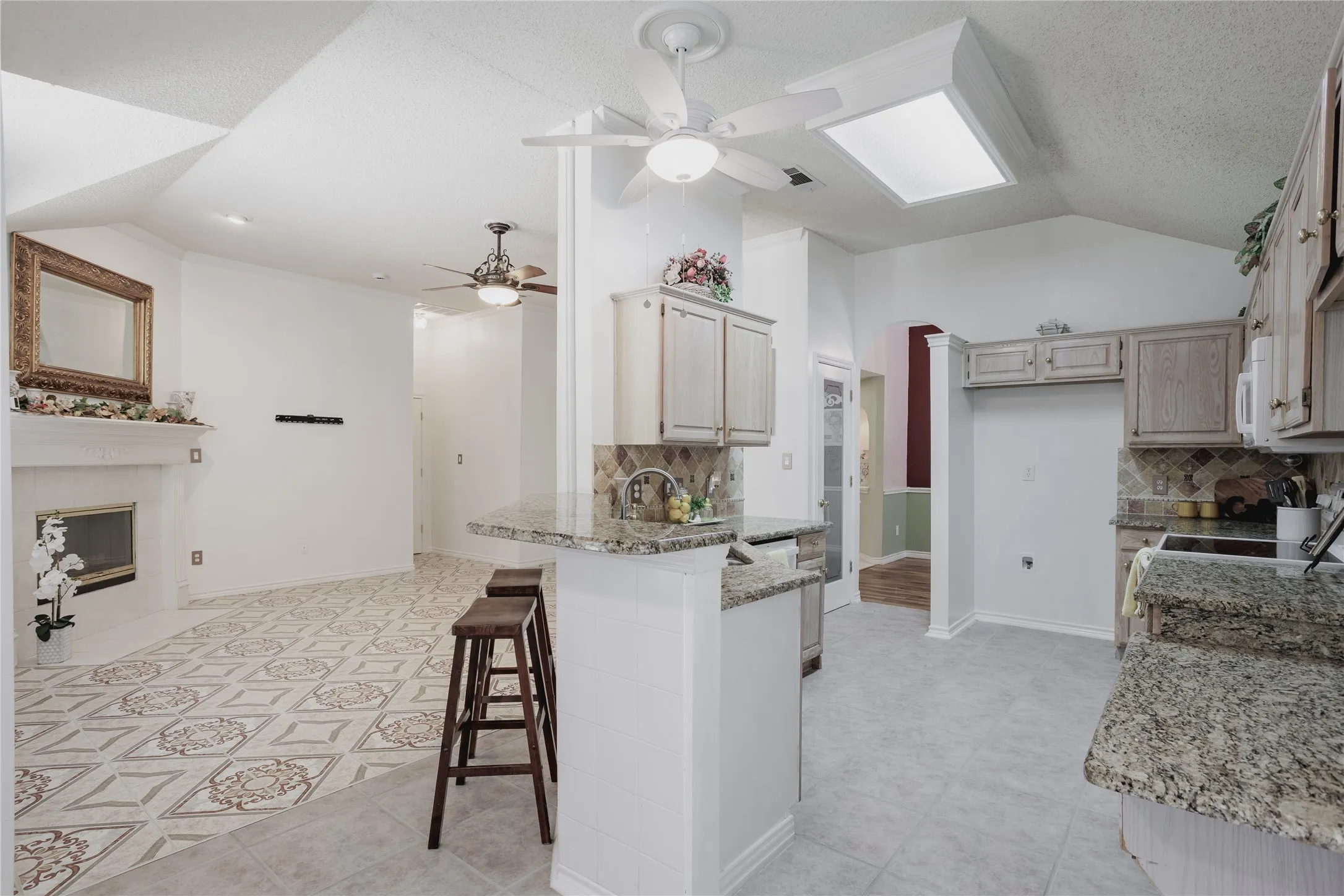 Bright, open kitchen and living area featuring vaulted ceilings with skylights, white cabinetry, granite countertops, a center island with seating, stainless steel appliances, and decorative tile flooring throughout the space