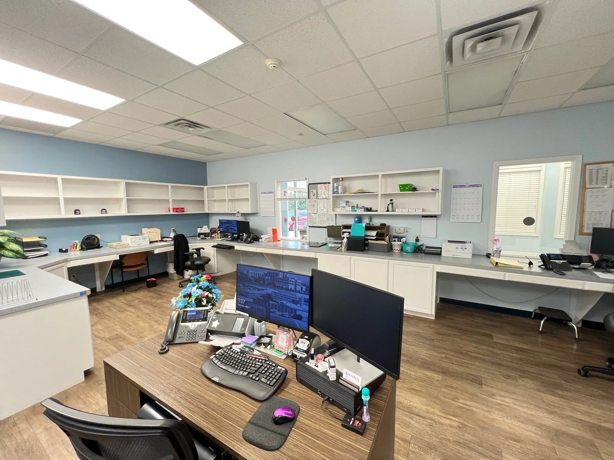 Spacious commercial office space featuring extensive built-in shelving, wood-finish flooring, ample countertop workspace, a dropped ceiling with recessed lighting, and a window for natural light