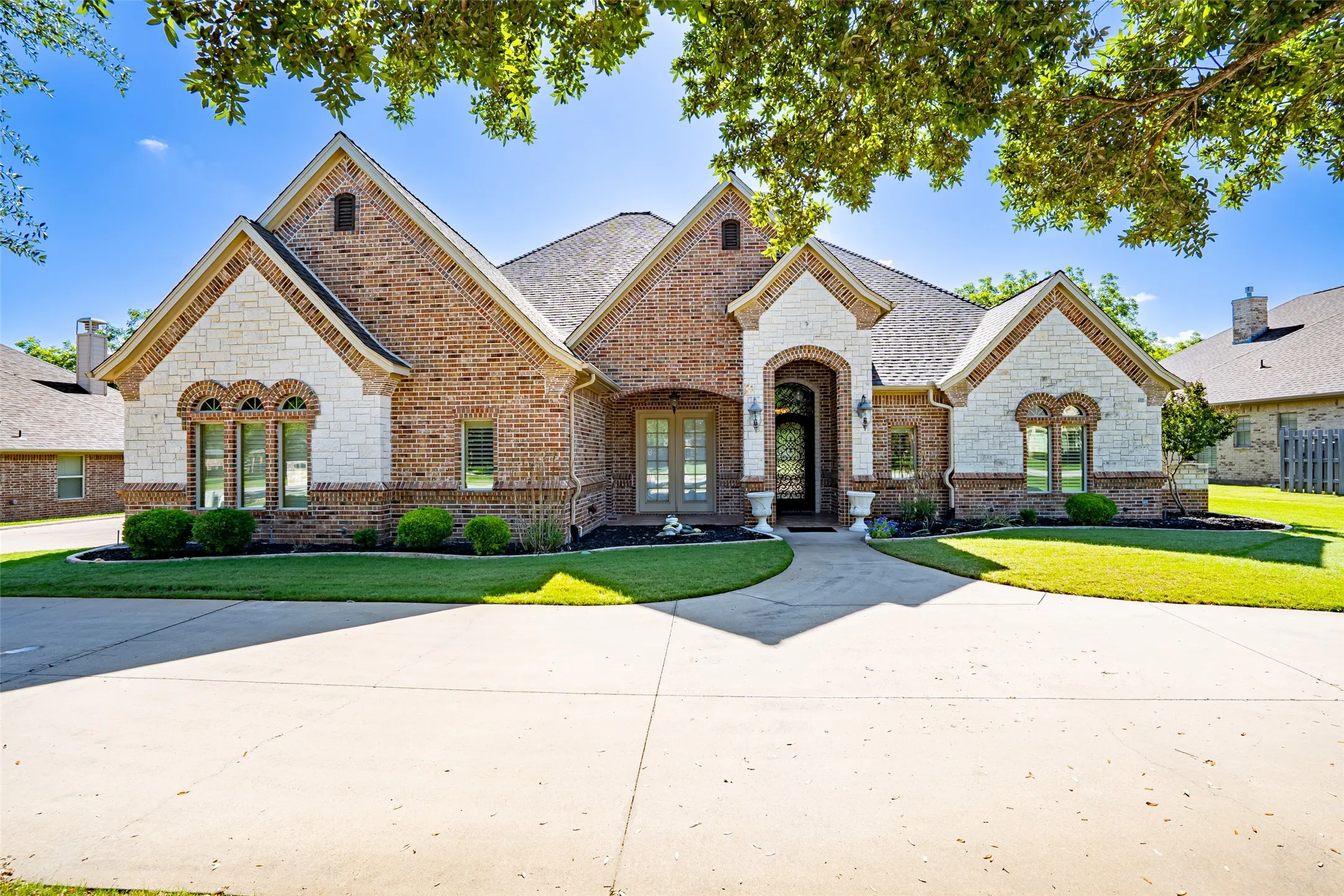Arched entry featuring an ornate wrought iron door, multi-tone brickwork, decorative brick pilasters, and a covered porch with tile flooring