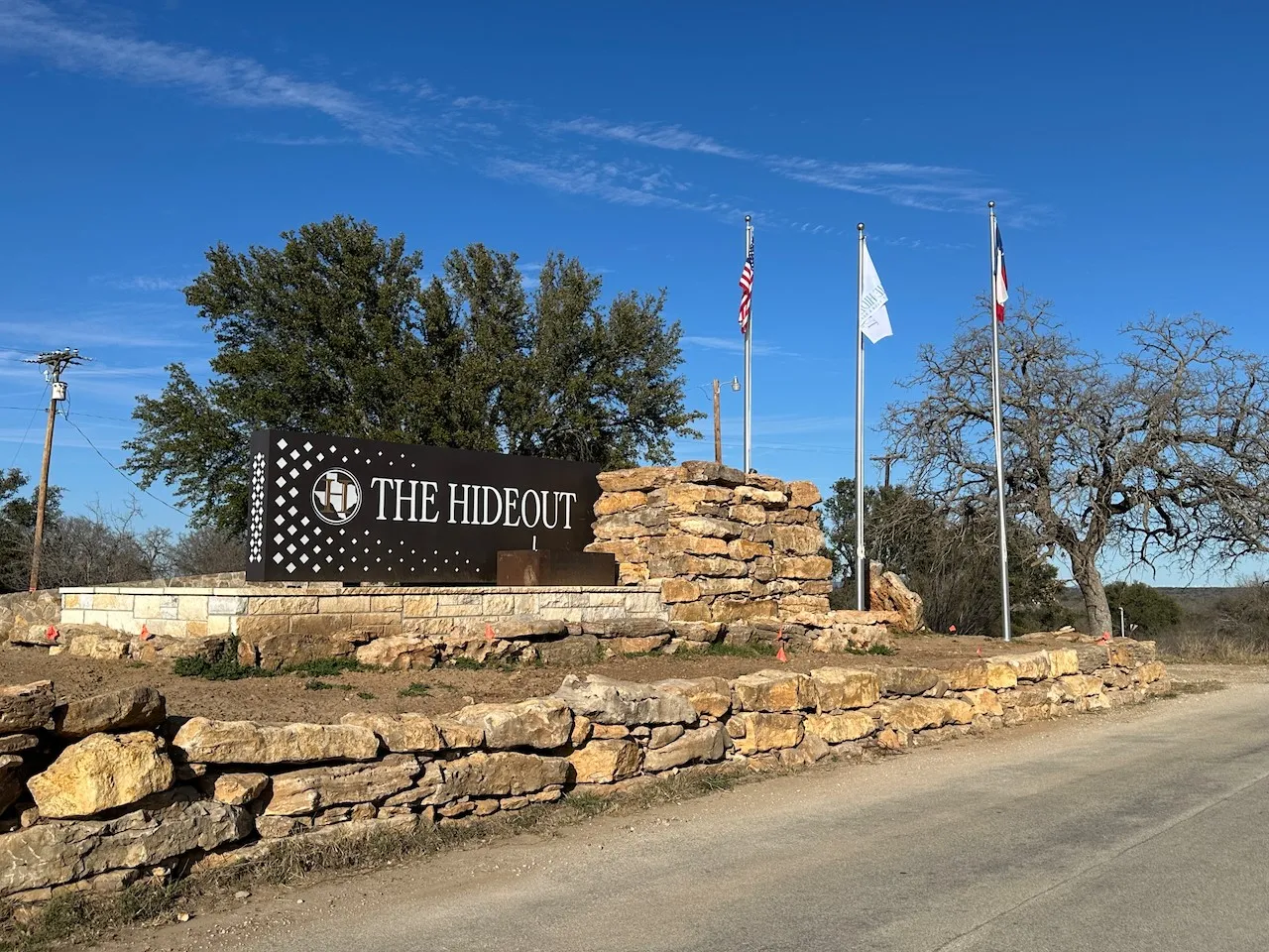 Custom stone entryway featuring a prominent "The Hideout" sign, multiple flagpoles with banners, and mature landscaping