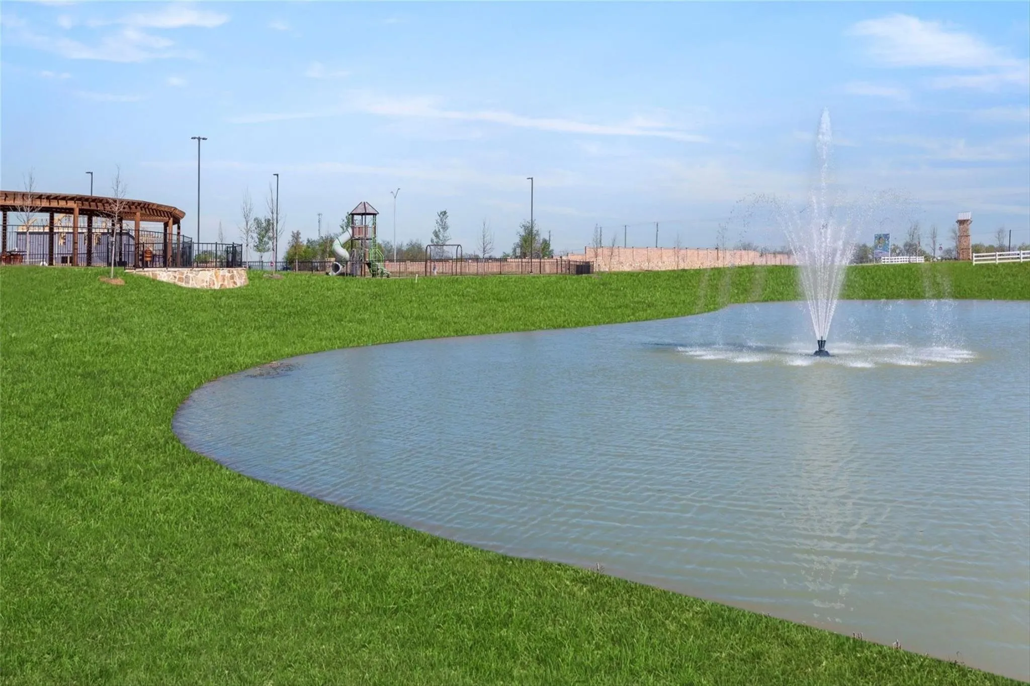 Manicured green space with a water feature, an expansive pond, a curved wooden pergola, and a children's play structure