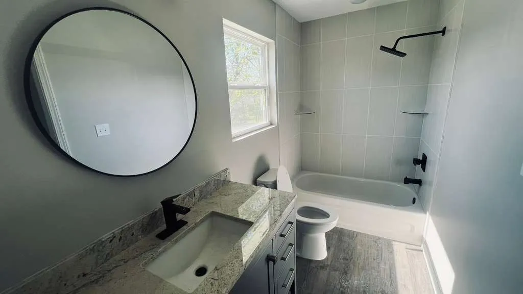 Bathroom featuring a modern circular mirror, granite-finish countertop, dark wood-finish vanity, and a tub-shower combination with light gray tile surround and matte black fixtures