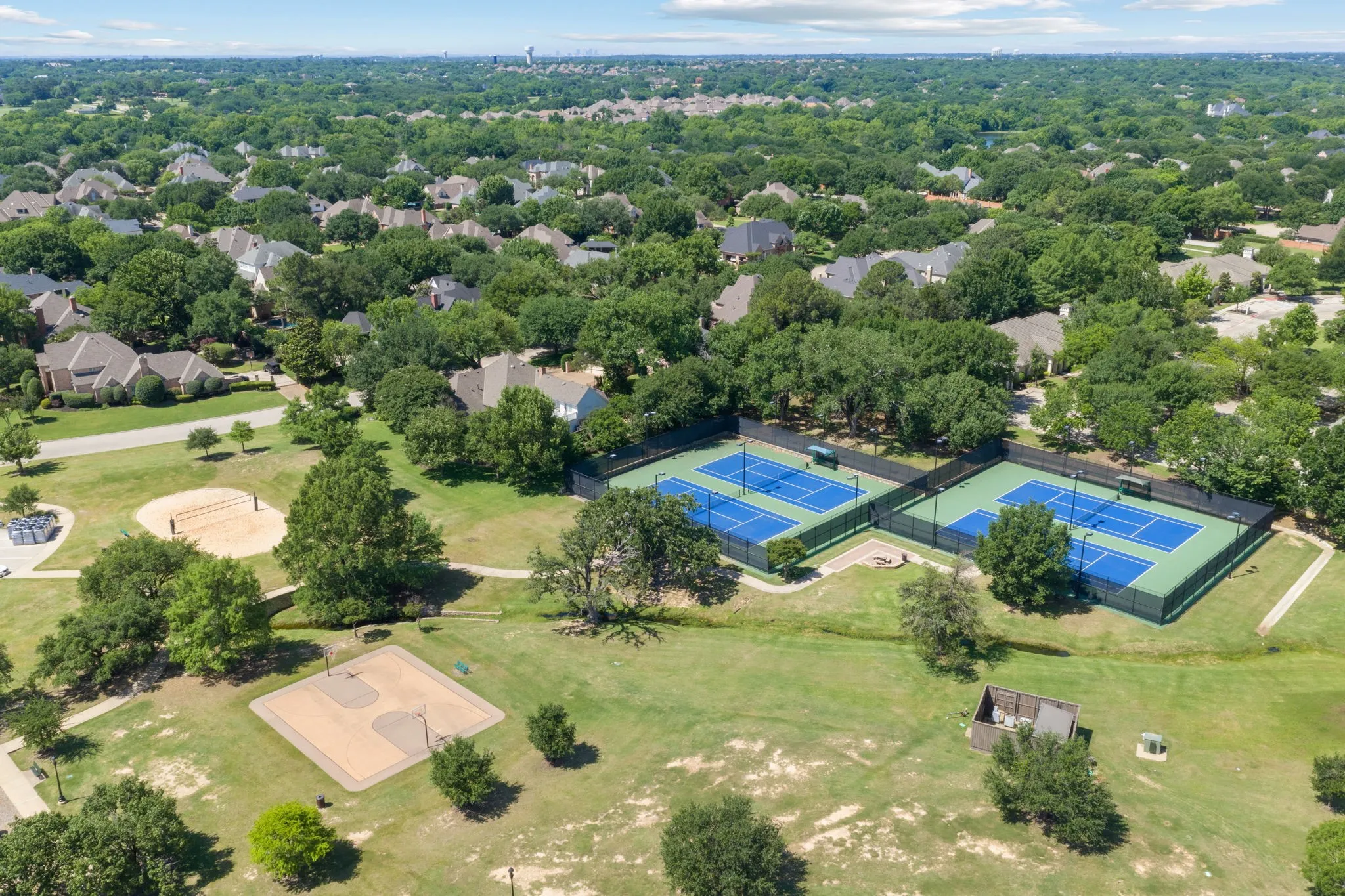 Two blue tennis courts with black fencing, a sand volleyball court, and an open-air basketball court set within a green space bordered by residential properties