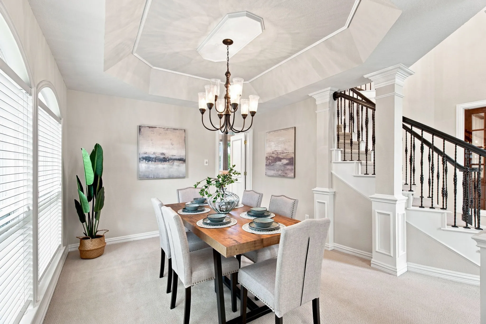 Recessed ceiling with decorative trim, arched window with horizontal blinds, chandelier lighting fixture, staircase with wrought iron balusters, and white paneled newel posts