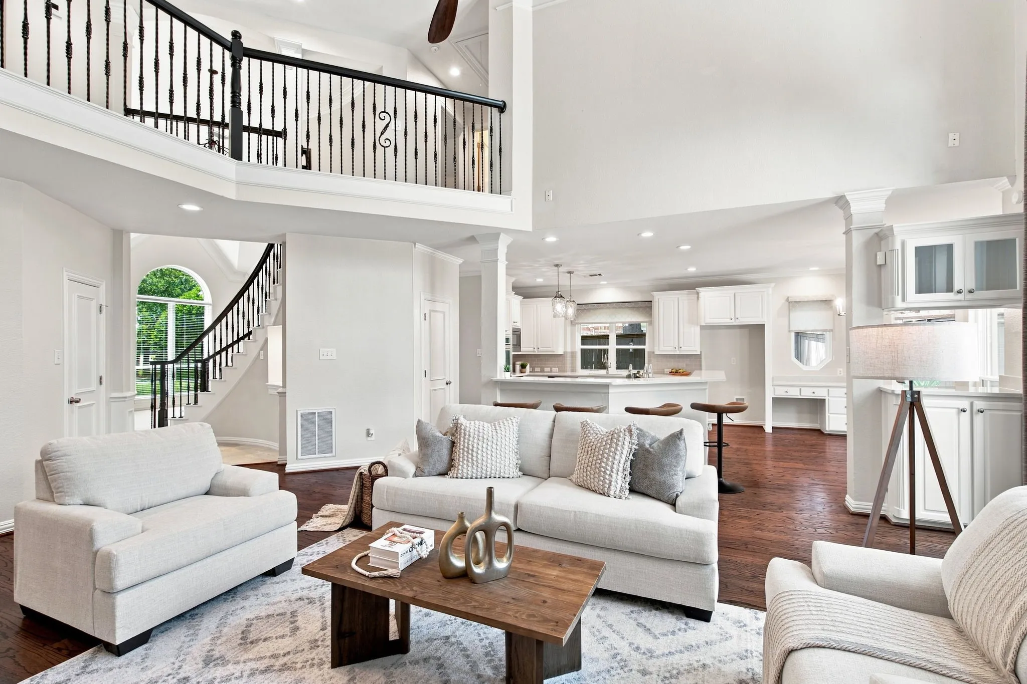 High vaulted ceilings, open concept floorplan, second-story landing with wrought iron railing, wood-finish flooring, and a bright kitchen with white cabinetry