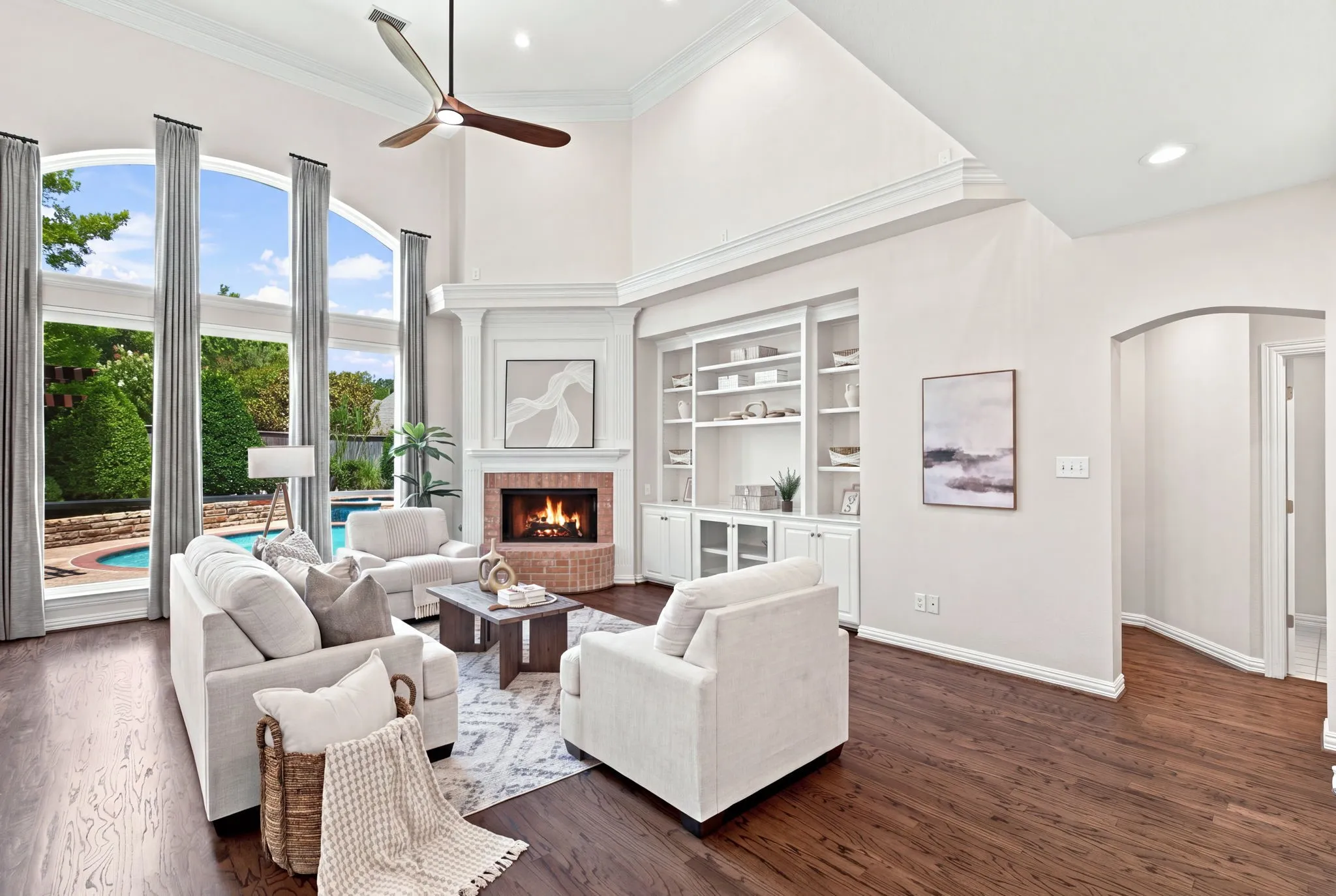 Vaulted ceiling with a ceiling fan, wood-finish flooring, brick-surround fireplace, built-in shelving, and large arched windows overlooking an outdoor swimming pool