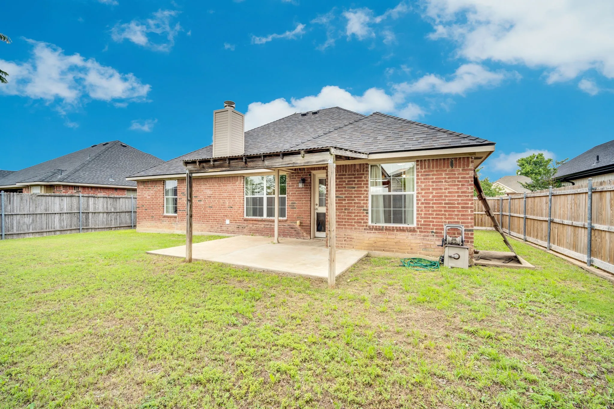 Expansive backyard featuring a covered concrete patio, brick exterior, dark shingle roof, and privacy fencing