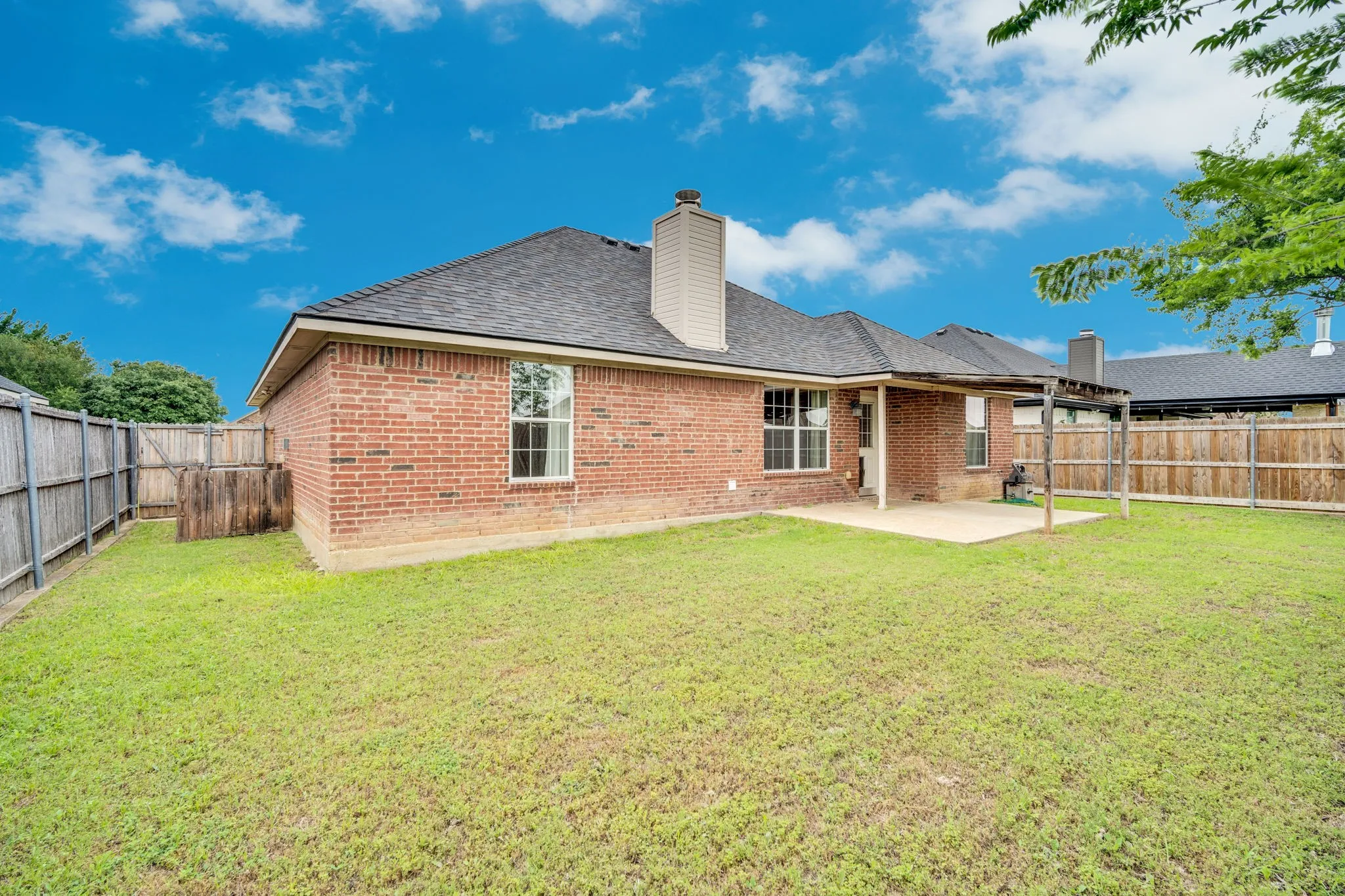 Expansive backyard featuring a red brick exterior, a covered patio, a concrete patio, a wooden pergola structure, and a perimeter fence