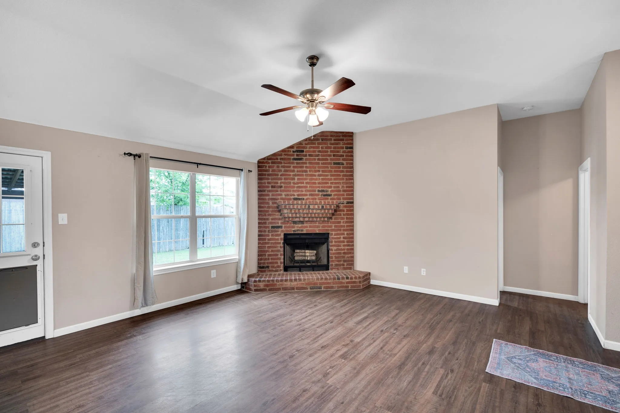 Spacious living area featuring a brick fireplace with hearth, vaulted ceiling, wood-finish flooring, and a ceiling fan