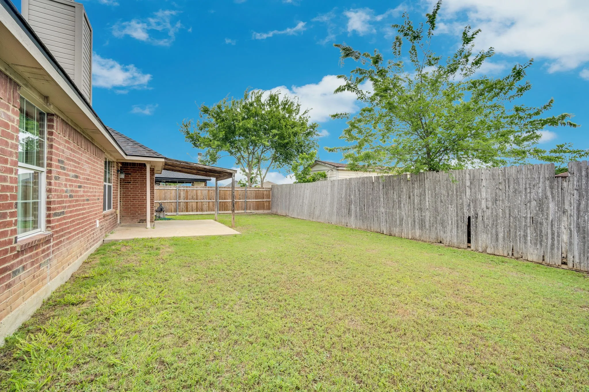 Expansive rear yard featuring green lawn, brick exterior, concrete patio, wood fence, and mature trees