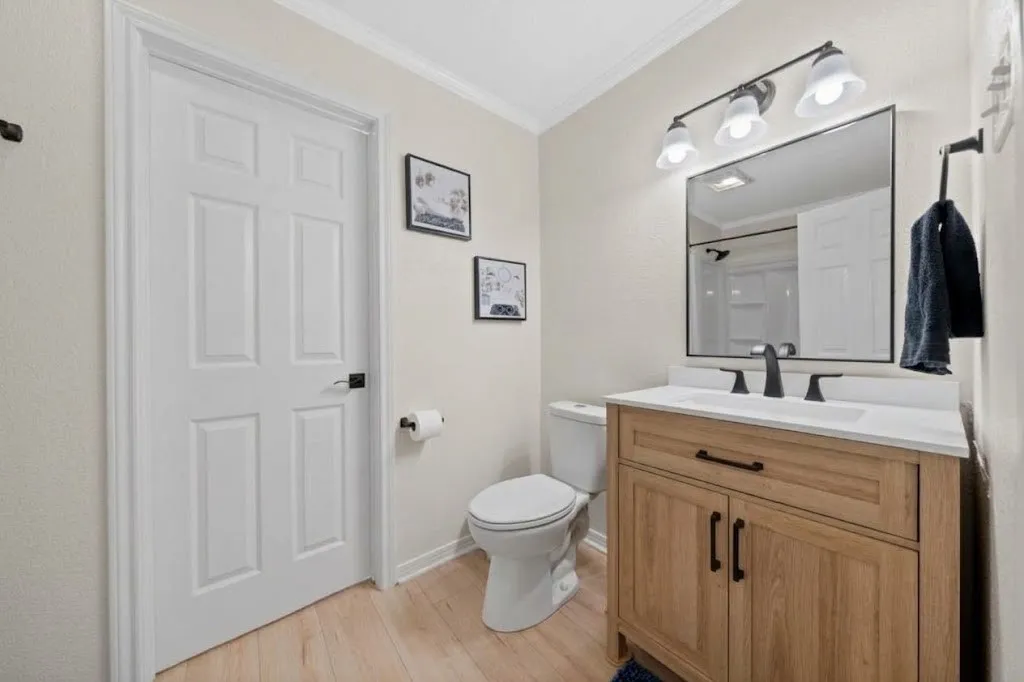 Light wood-finish flooring, white vanity with quartz countertop, black-framed mirror, and three-light sconce