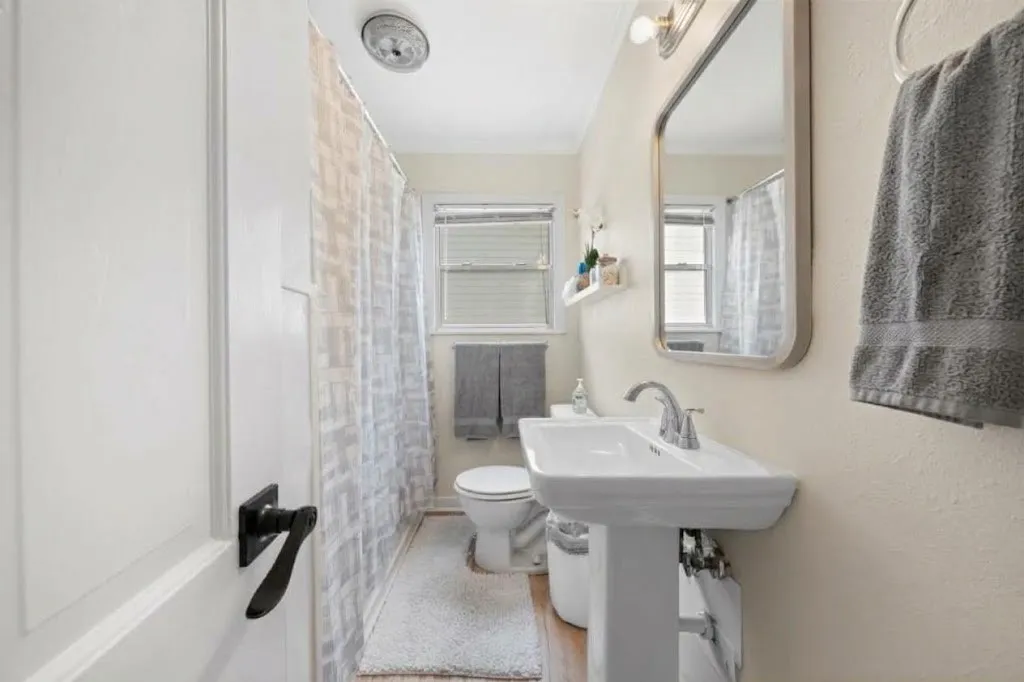 Pedestal sink, wall-mounted mirror, brushed nickel fixtures, window with blinds, and wood-finish flooring