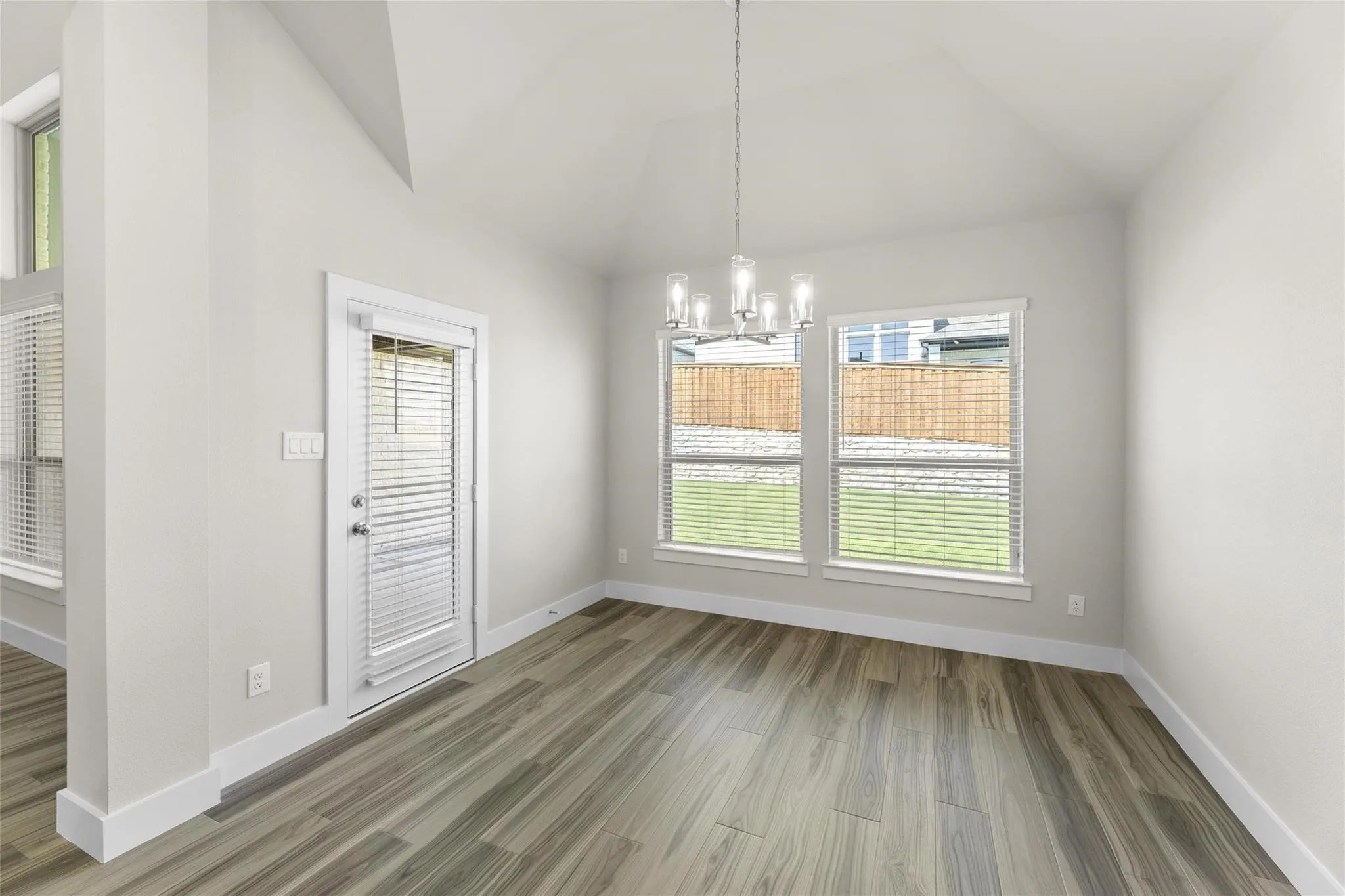 Spacious room featuring vaulted ceilings, wood-finish flooring, a contemporary chandelier, and multiple windows with blinds