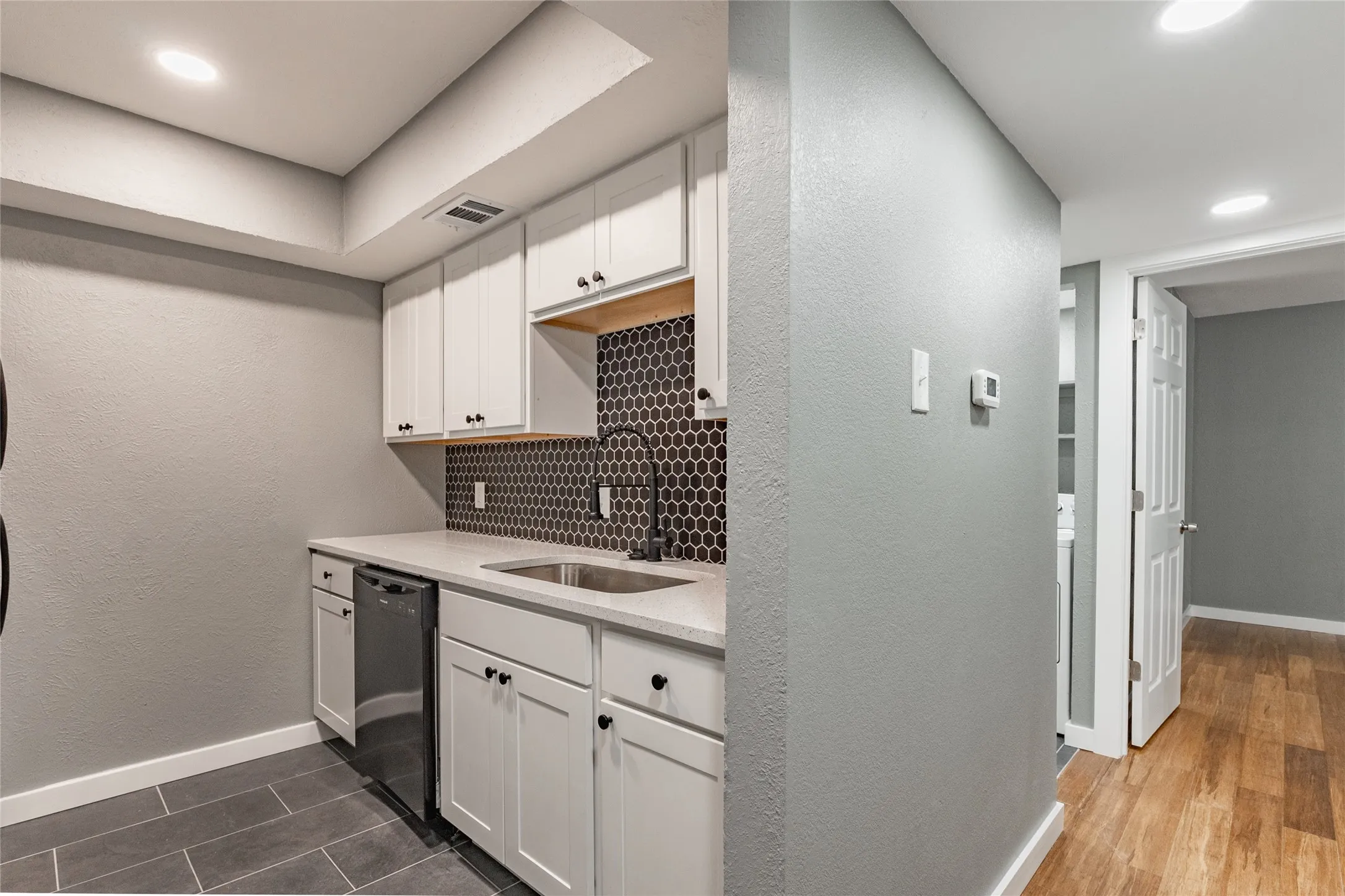 Kitchenette featuring white cabinetry, a black honeycomb tile backsplash, light countertops, a stainless steel sink with a matte black faucet, and a built-in dishwasher