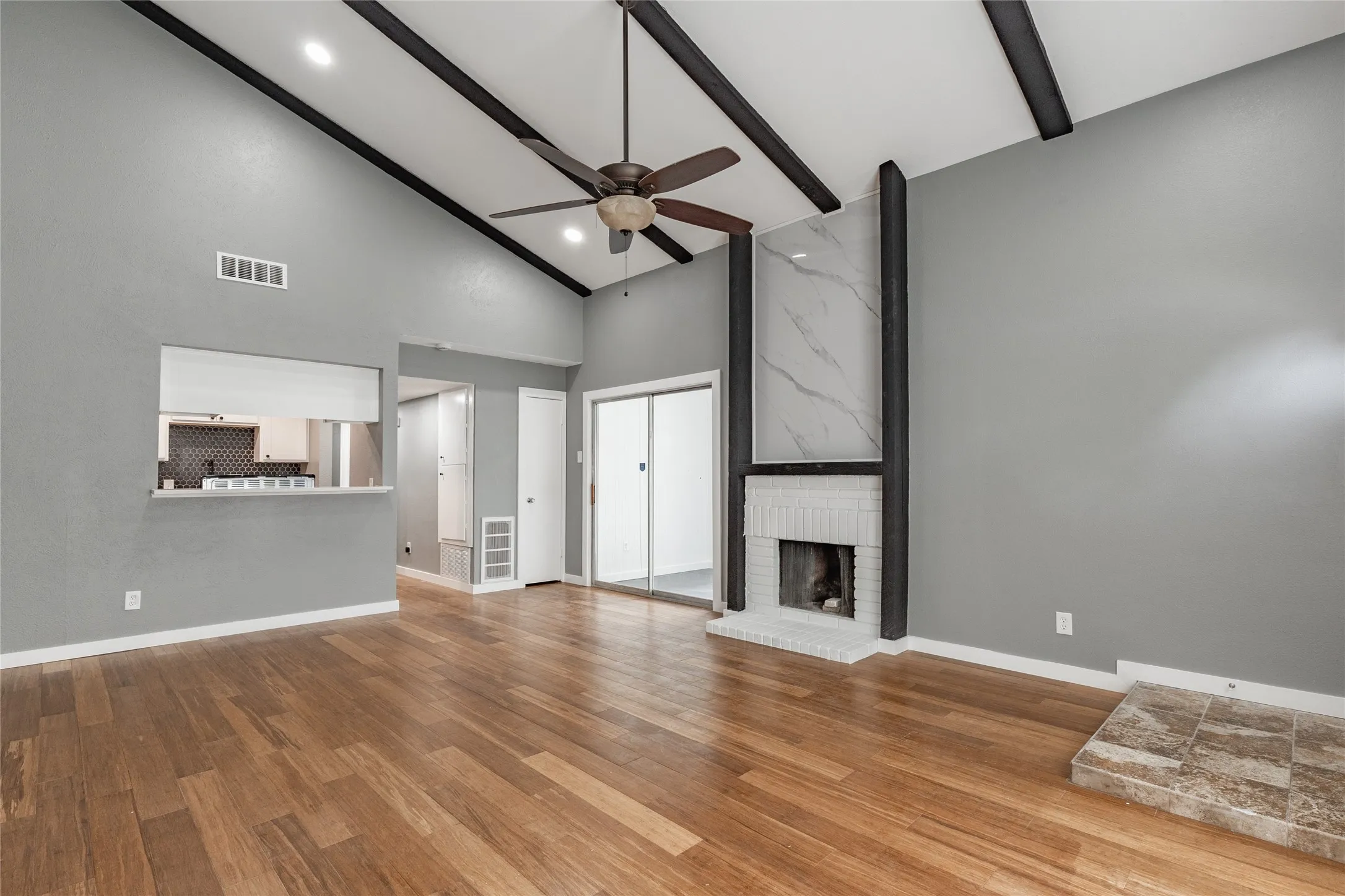 Spacious room featuring vaulted ceilings with exposed beams, wood-finish flooring, and a white brick fireplace with a marble-look surround