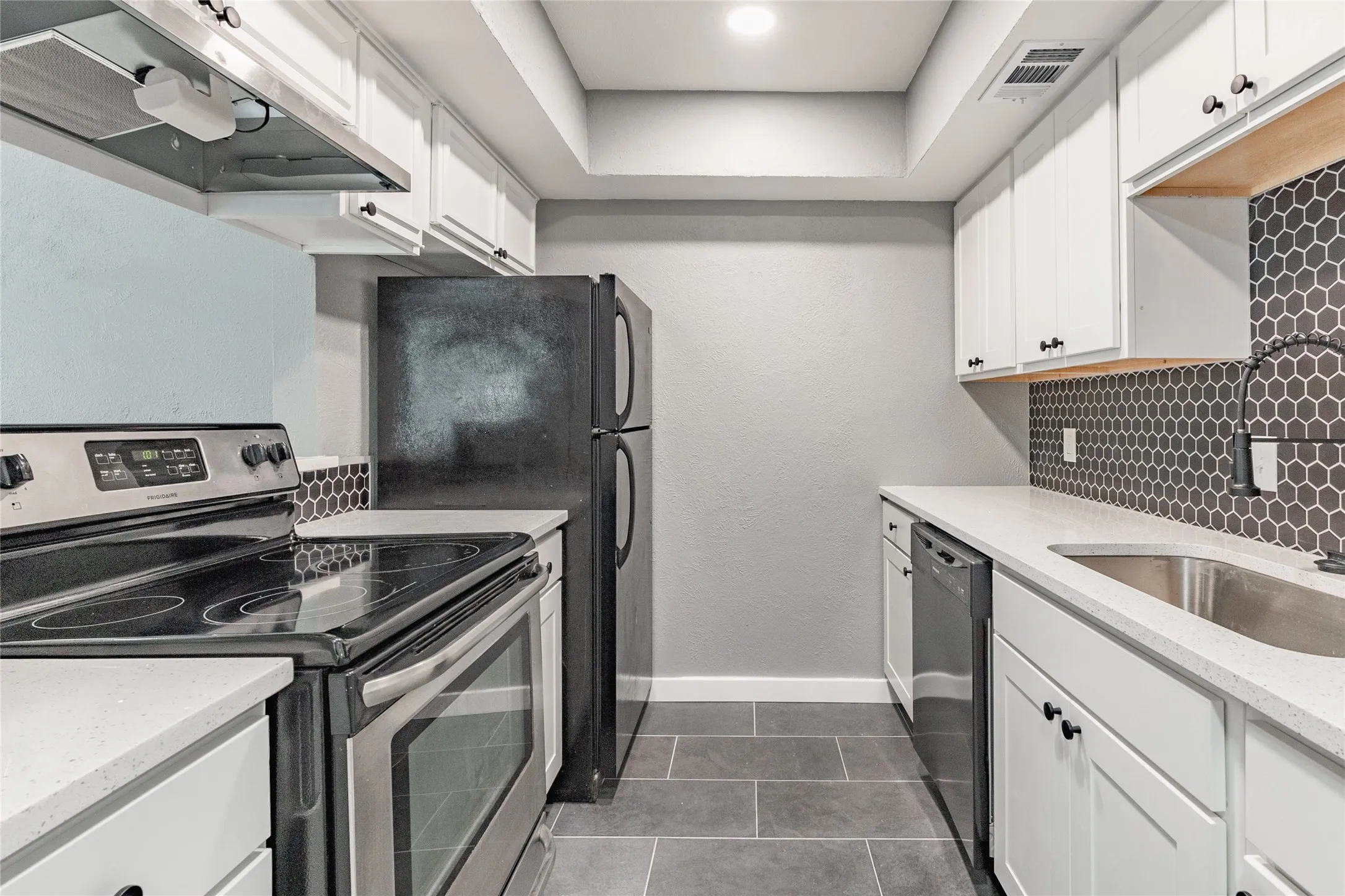 Galley kitchen featuring white shaker cabinetry, light-toned countertops, a full suite of black and stainless steel appliances, a dark hexagonal tile backsplash, and dark tile flooring