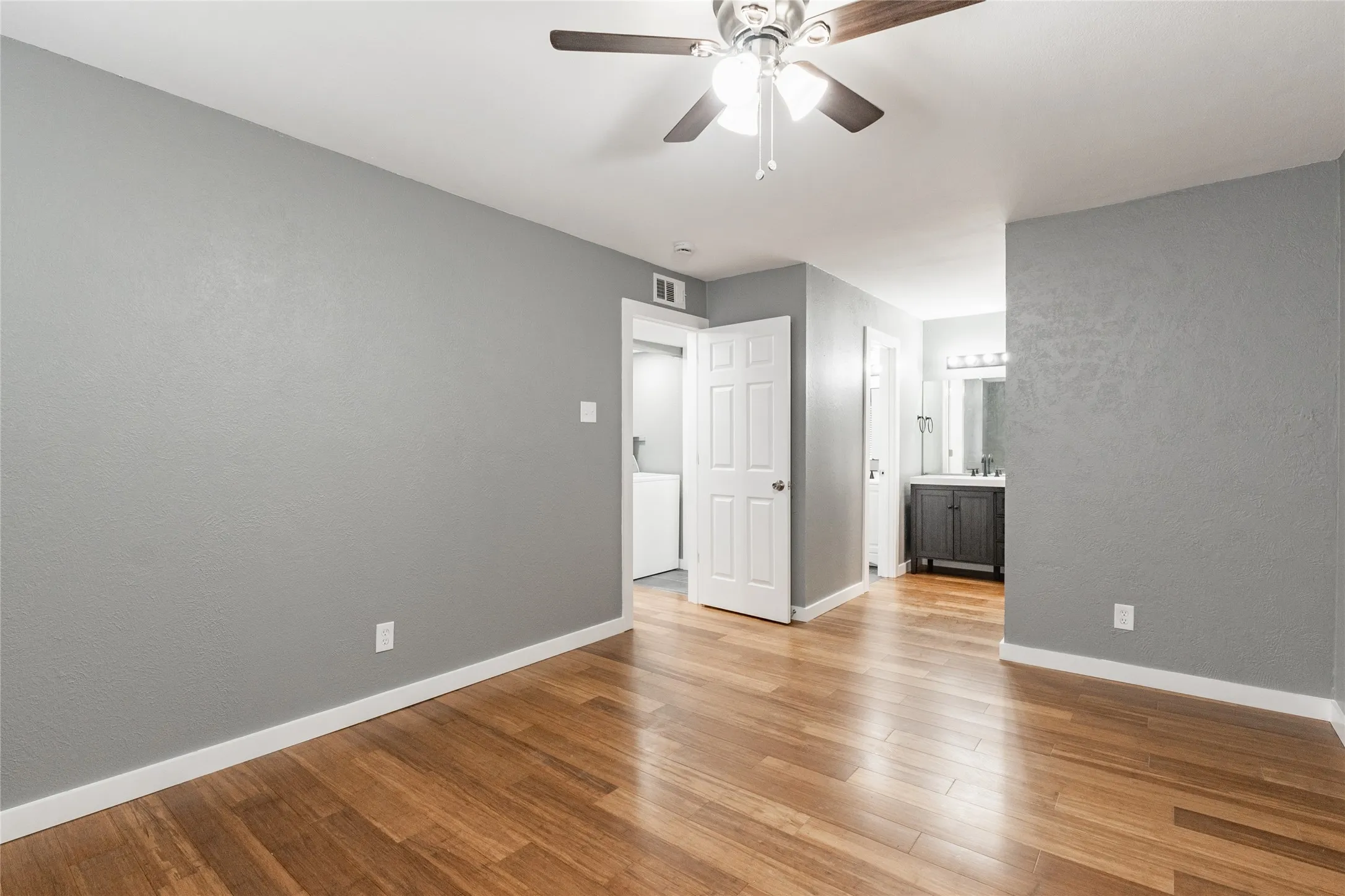 Spacious room featuring wood-finish flooring, light gray walls, a ceiling fan with integrated lighting, and white trim