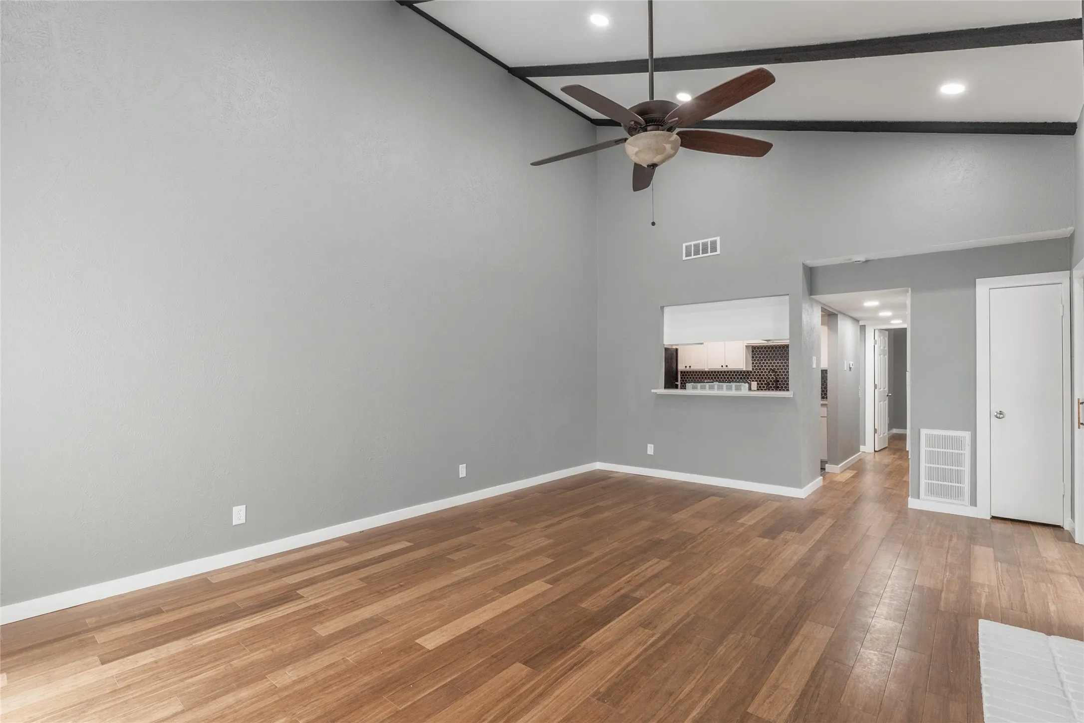 Spacious living area featuring high vaulted ceilings with exposed beams, wood-finish flooring, a ceiling fan, and a pass-through window to the kitchen