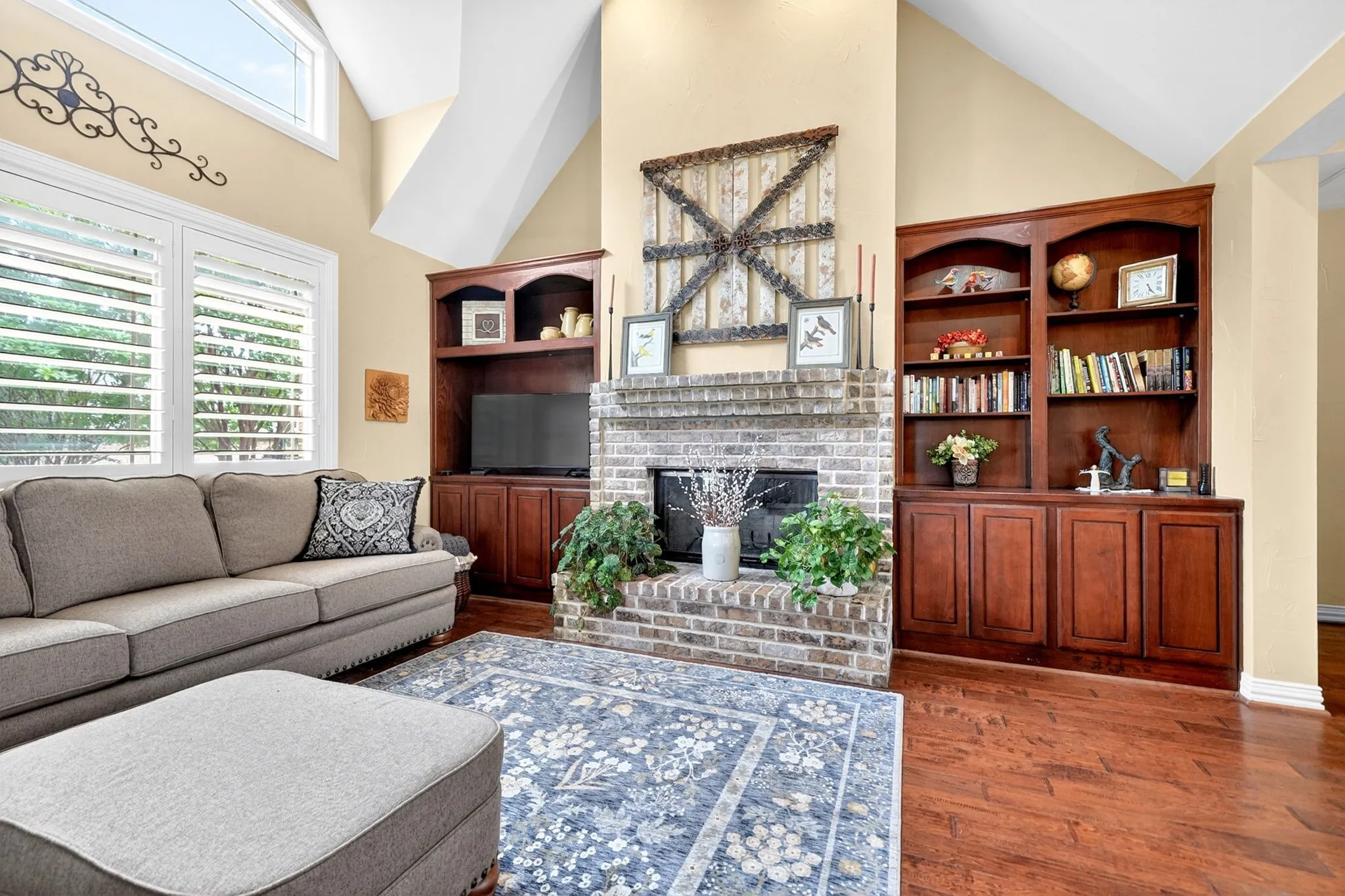Living area featuring a brick fireplace, built-in shelving, rich wood-finish flooring, plantation shutters, and vaulted ceilings