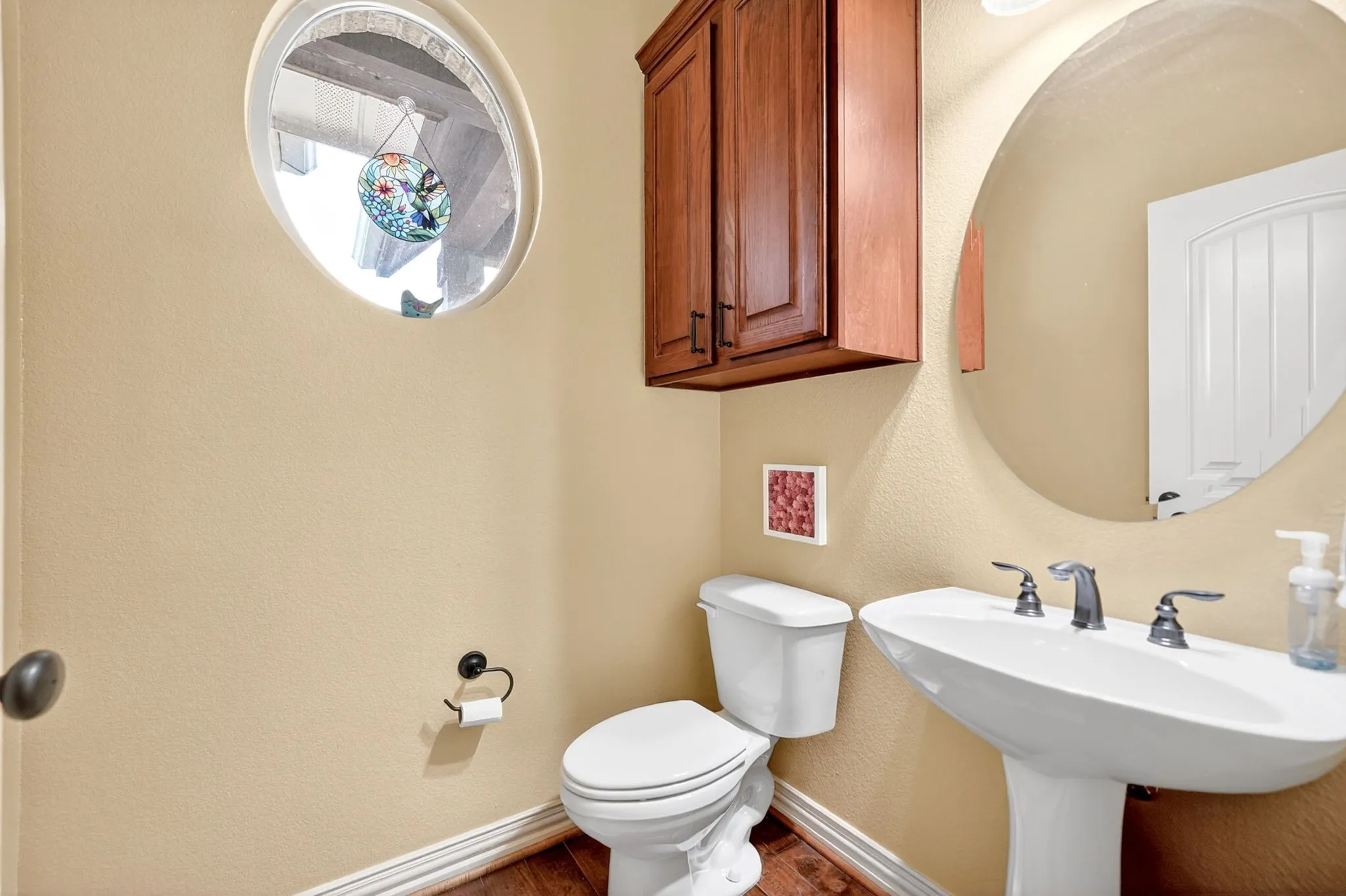 Pedestal sink with chrome fixtures, wood-finish flooring, and a distinctive oval window