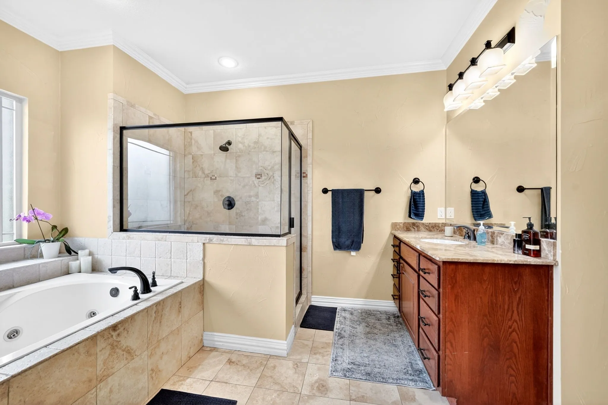 Spacious bathroom featuring a glass-enclosed shower with multiple showerheads, a deep soaking tub with matte black fixtures, a dual-sink vanity with a stone-finish countertop, wood-finish cabinetry, and recessed lighting