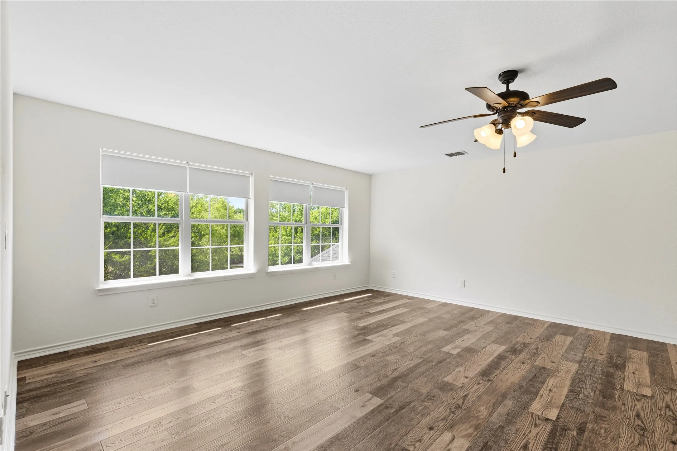 Spacious room featuring wood-finish flooring, multiple windows with roller shades, and a ceiling fan with integrated lighting