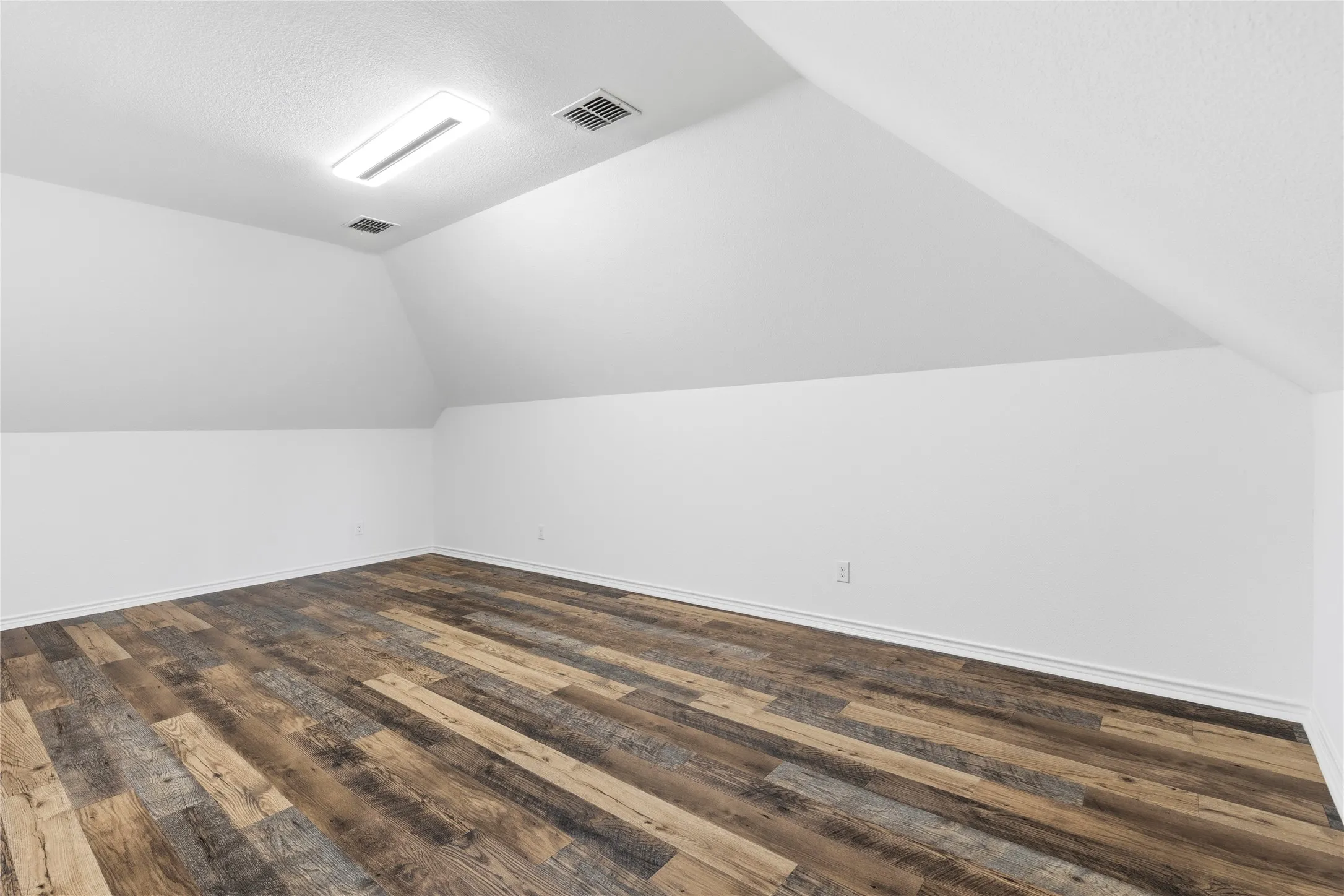 Spacious room featuring sloped ceilings, white walls, wood-finish flooring, and a flush-mount ceiling light fixture