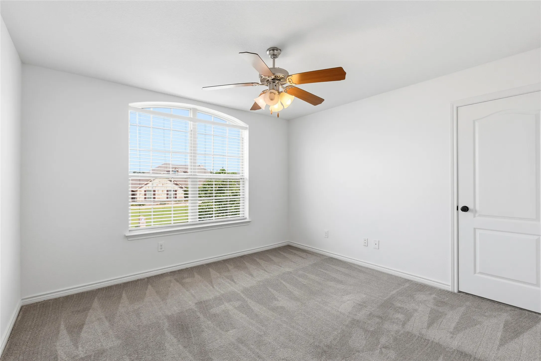 Spacious room featuring light gray patterned carpeting, a large arched window with horizontal blinds, and a ceiling fan with integrated lighting