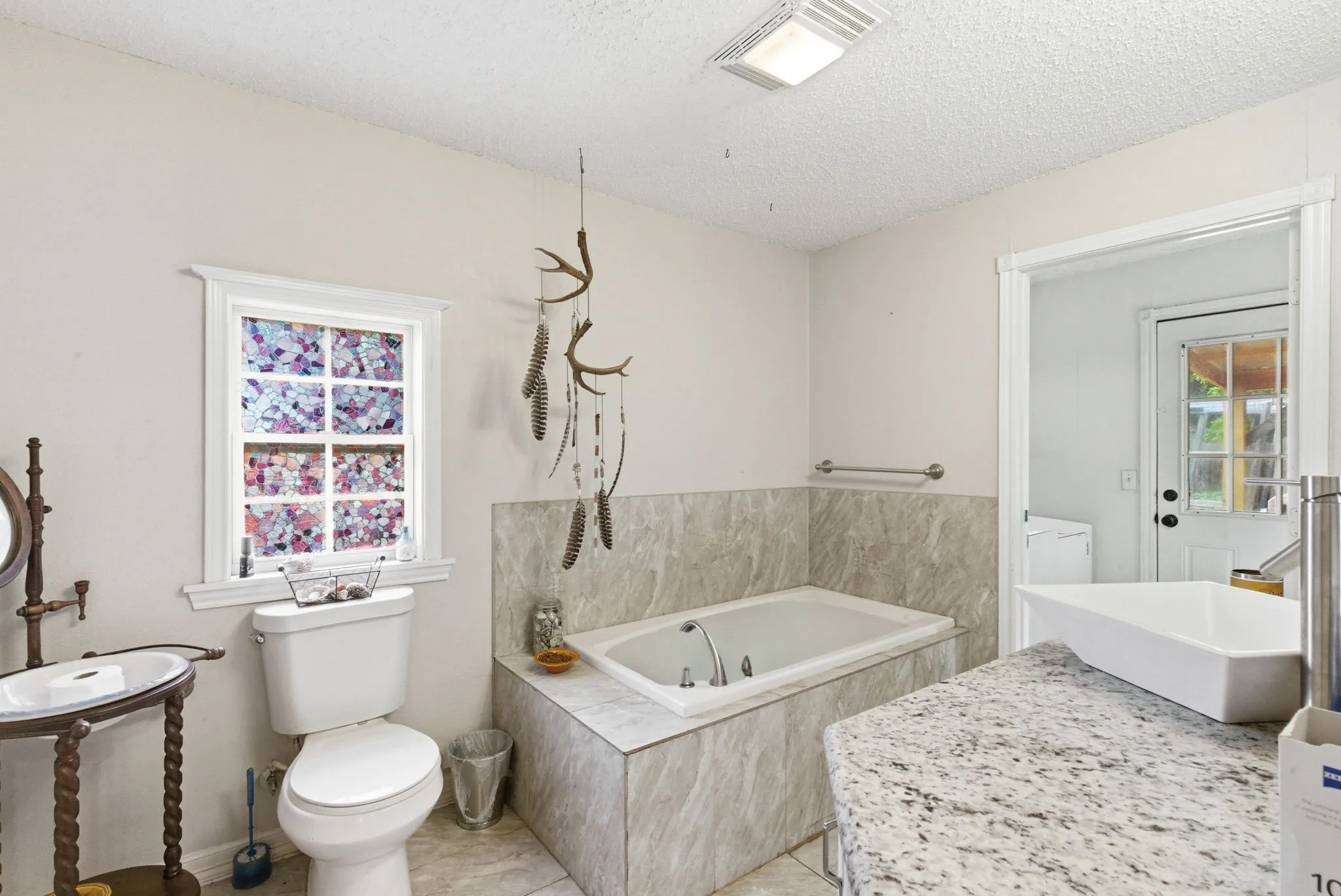 Bathroom featuring a drop-in bathtub with tiled surround, a white toilet, and a vanity with a rectangular vessel sink and speckled countertop